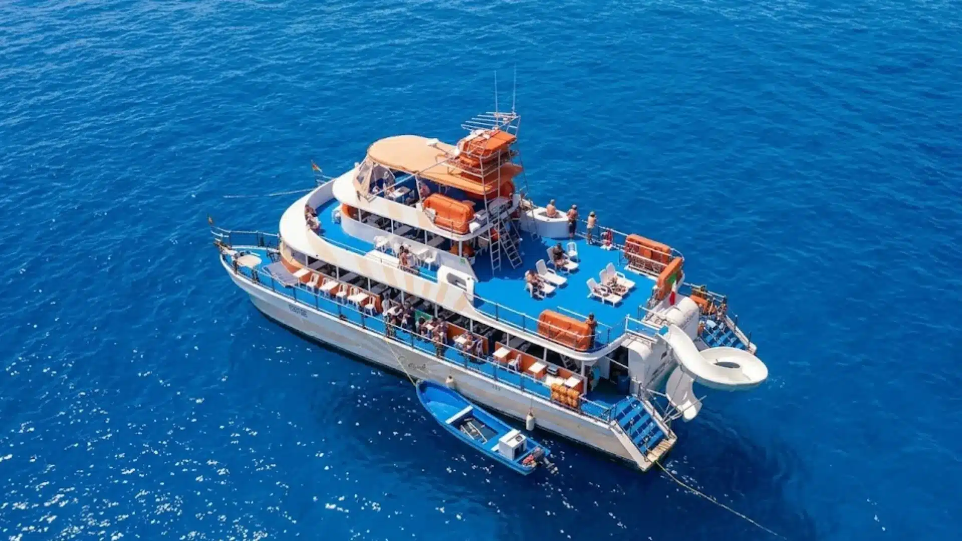 Aerial view of boat party with people relaxing on deck surrounded by clear blue ocean and bright sunlight