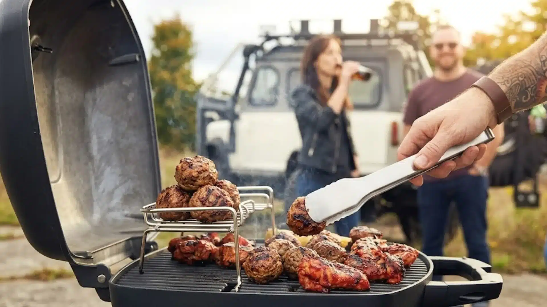 Hand grilling meatballs and chicken on barbecue with friends and car in outdoor casual gathering setting