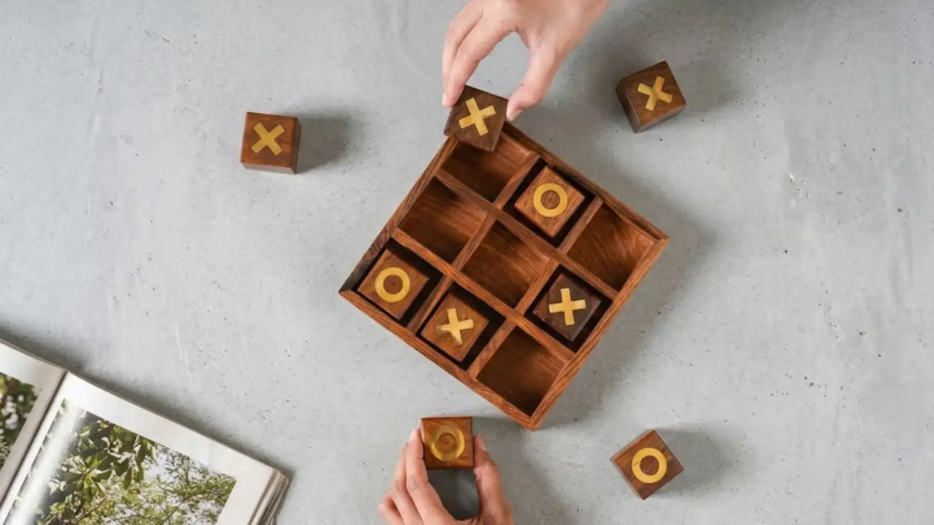 Hands placing wooden tic tac toe pieces on board game with X and O tiles on light surface