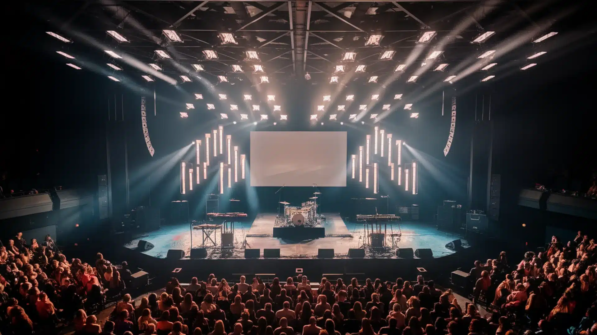Indoor concert venue with bright stage lights, band setup, and large audience seated for live performance