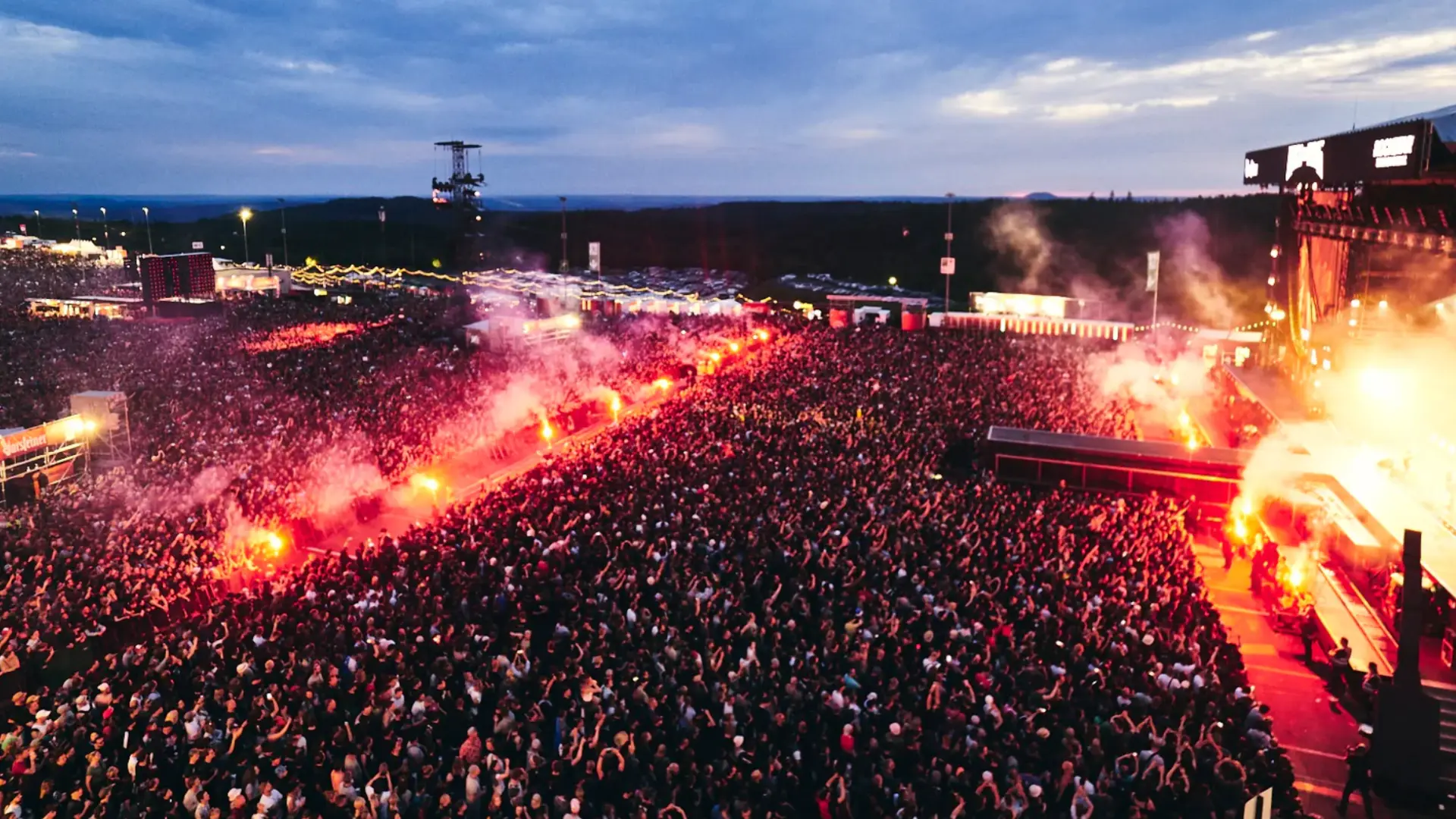 Massive outdoor concert crowd at dusk with flares lighting up stage and thousands of fans packed tightly together