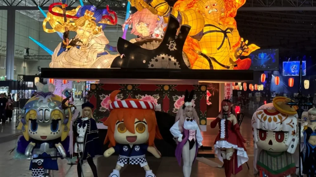 Anime convention display with colorful Fate characters, mascots, and cosplayers posing in front of festival-style stage
