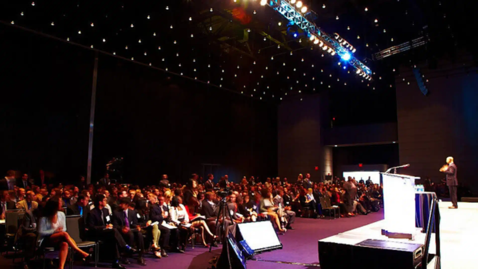 Speaker addressing large audience in conference hall with stage lighting and attendees seated in rows