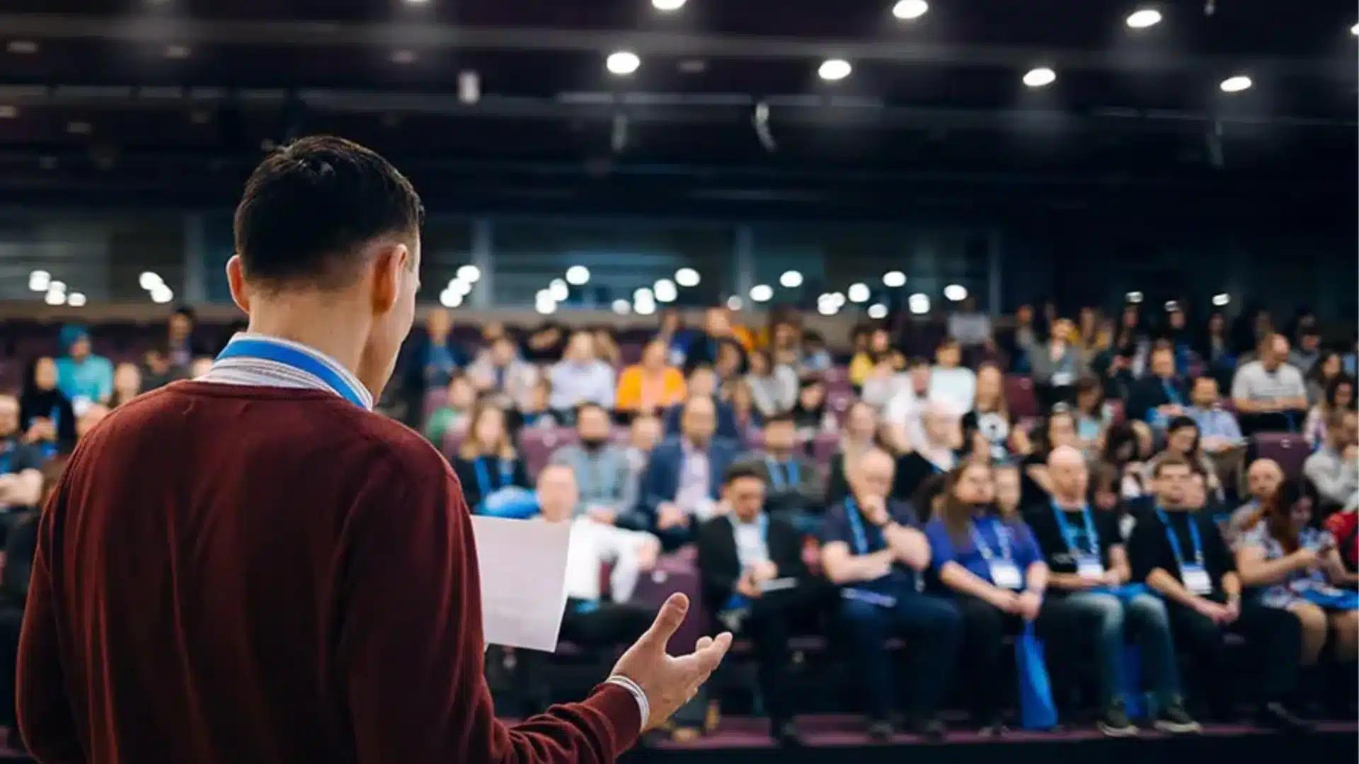 Speaker presenting to a large audience at a conference or seminar with attendees seated and focused on stage