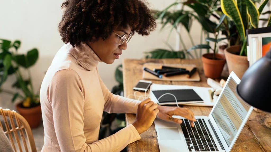 A woman with curly hair and glasses works at a wooden desk with a laptop, notebook, phone, and pen, surrounded by potted plants in a well-lit room, preparing video conference tips