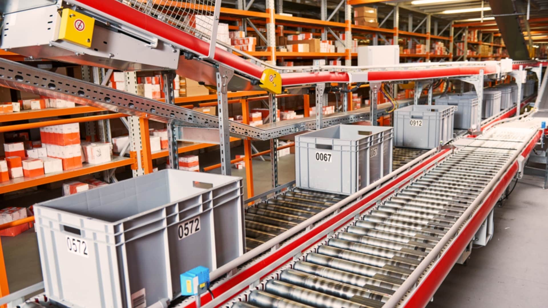 Plastic bins labeled with numbers move along a roller conveyor in a warehouse, illustrating the benefits of automated warehouse systems, with shelves of boxes and packages in the background highli