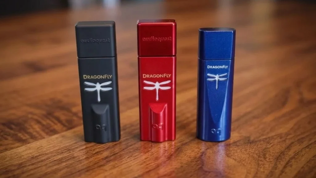 AudioQuest DragonFly Black, Red, and Cobalt USB DAC headphone amplifiers placed on a wooden table showing three devices