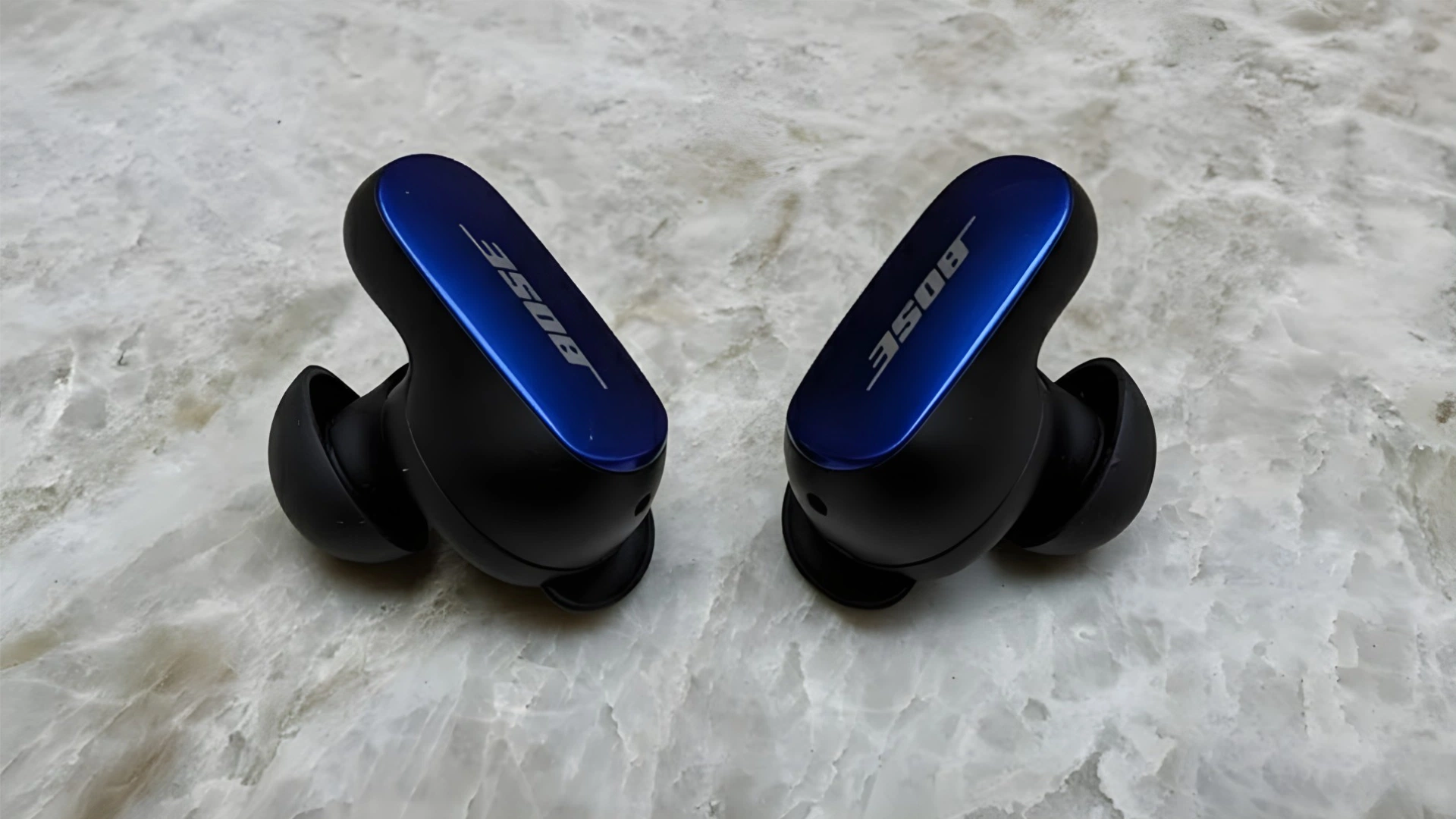 Bose QuietComfort Ultra earbuds in blue and black placed on marble surface close up