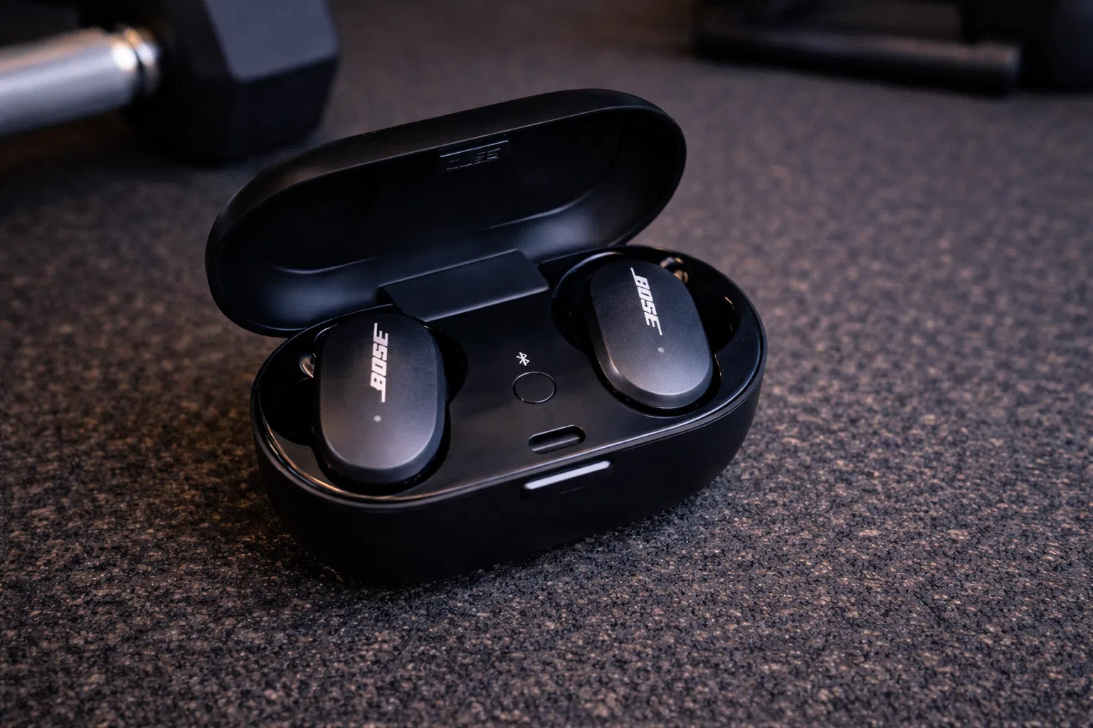 Bose QuietComfort true wireless earbuds in black charging case on gym floor near dumbbell