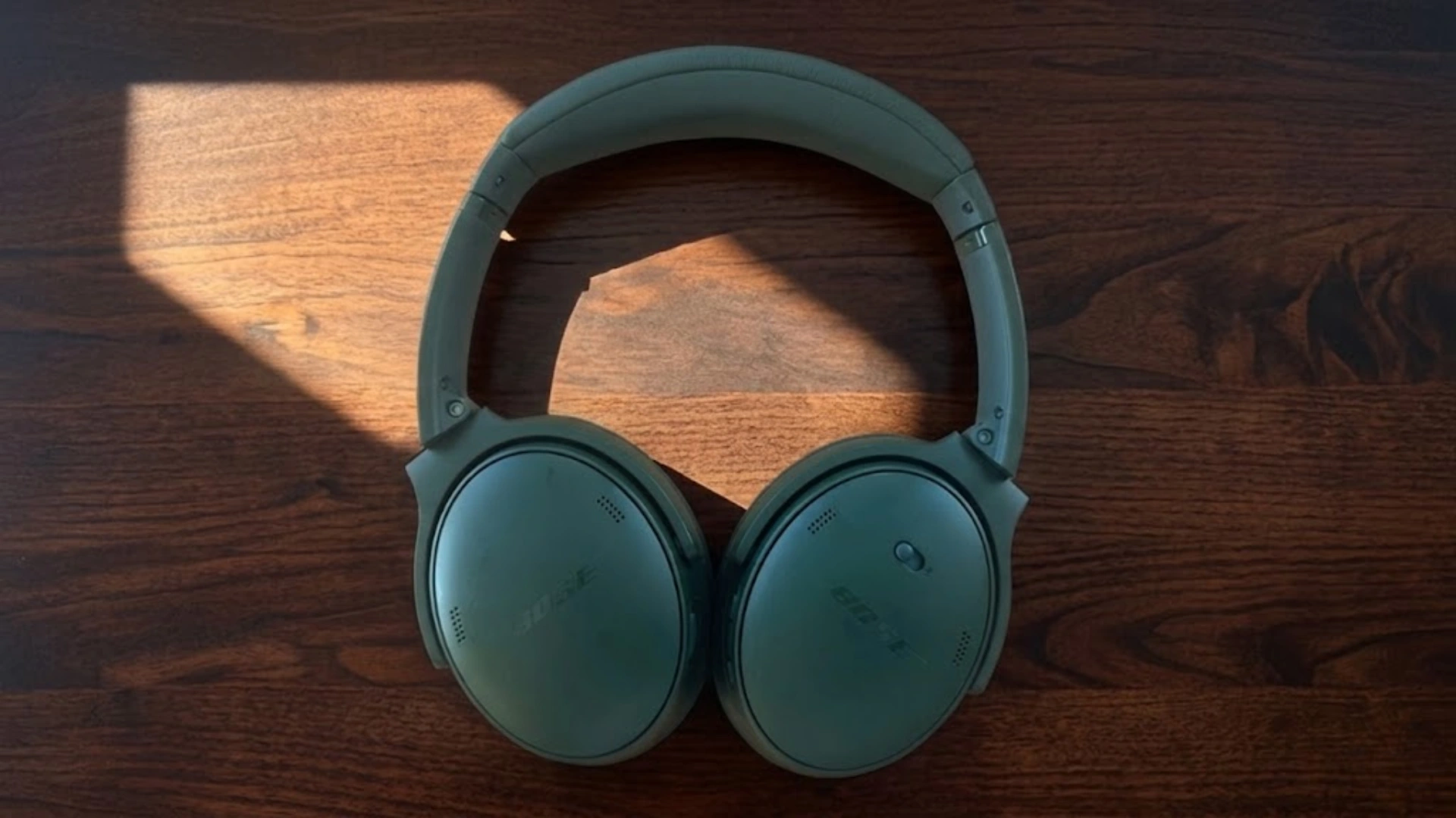 Bose quietcomfort over ear headphones placed on wooden table with natural sunlight highlighting the ear cups and headband design