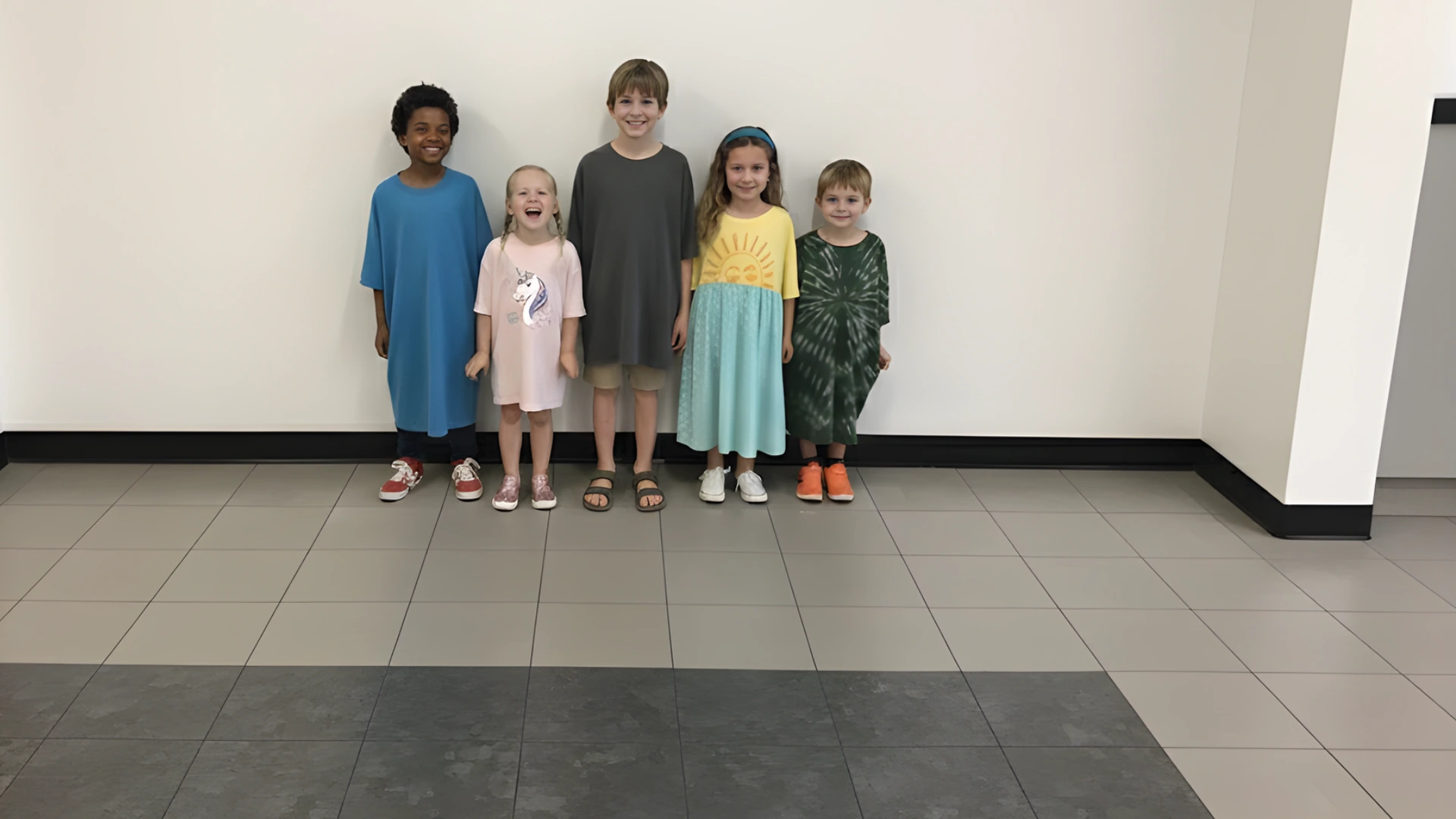 Five children wearing oversized T shirts and dresses standing together against classroom wall for spirit day