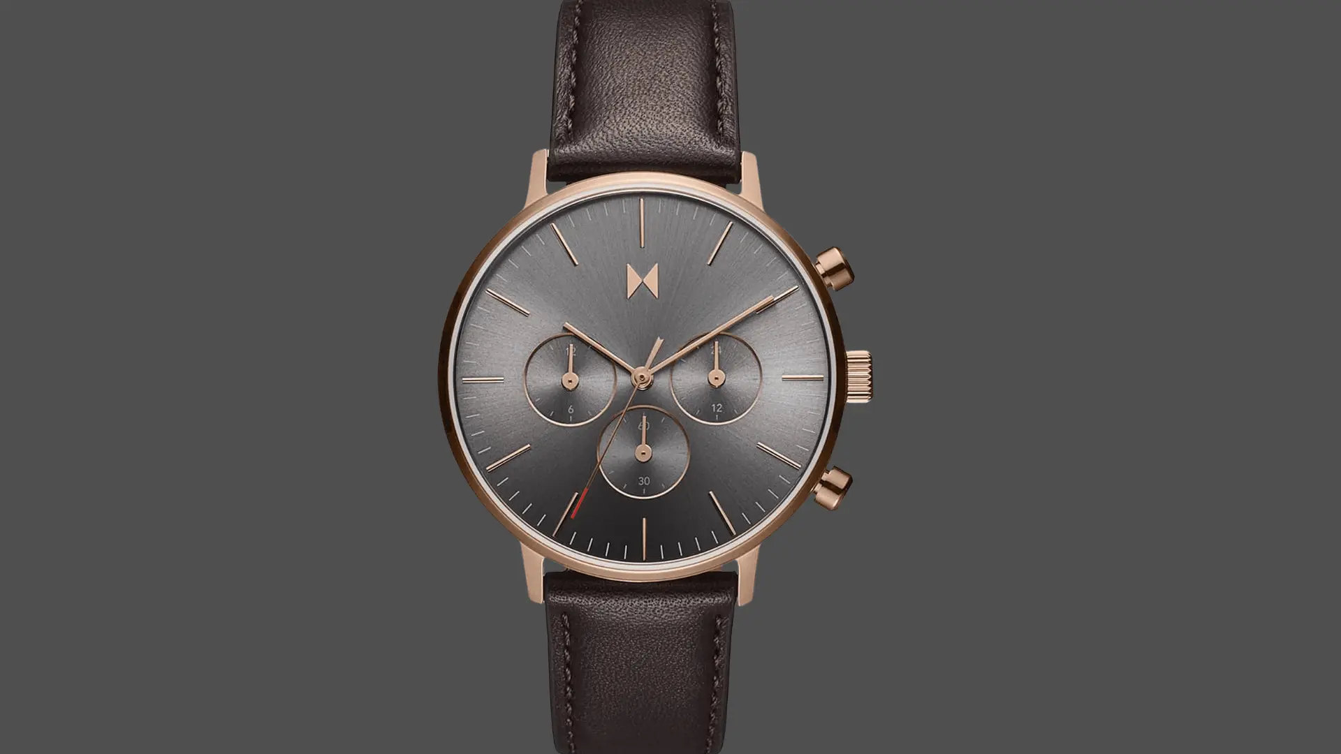 MVMT chronograph watch with gray dial, rose gold case, and dark leather strap on a simple gray background