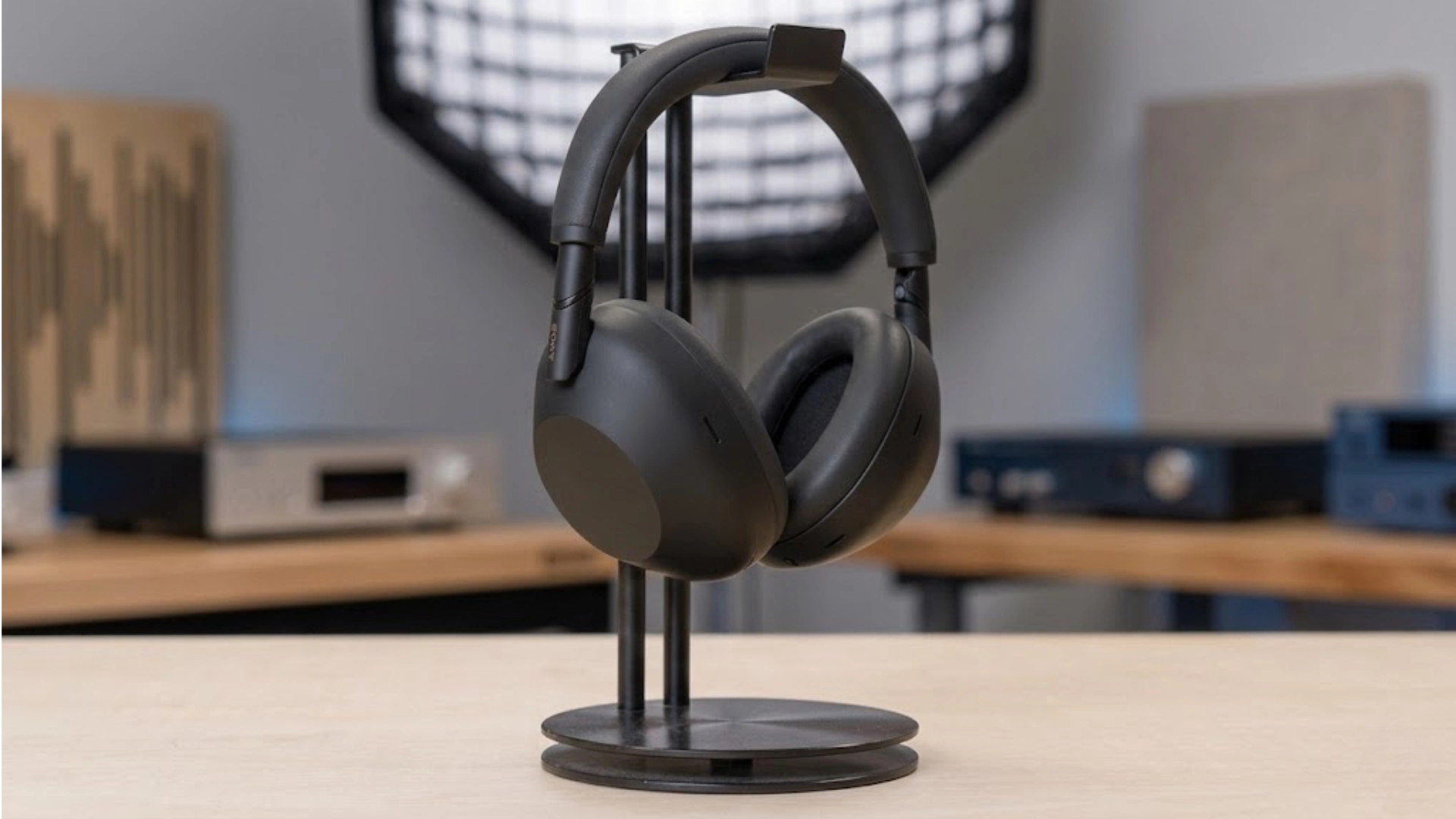 Sony WH-1000XM6 wireless noise cancelling headphones placed on a stand in a desk setup with audio equipment in background