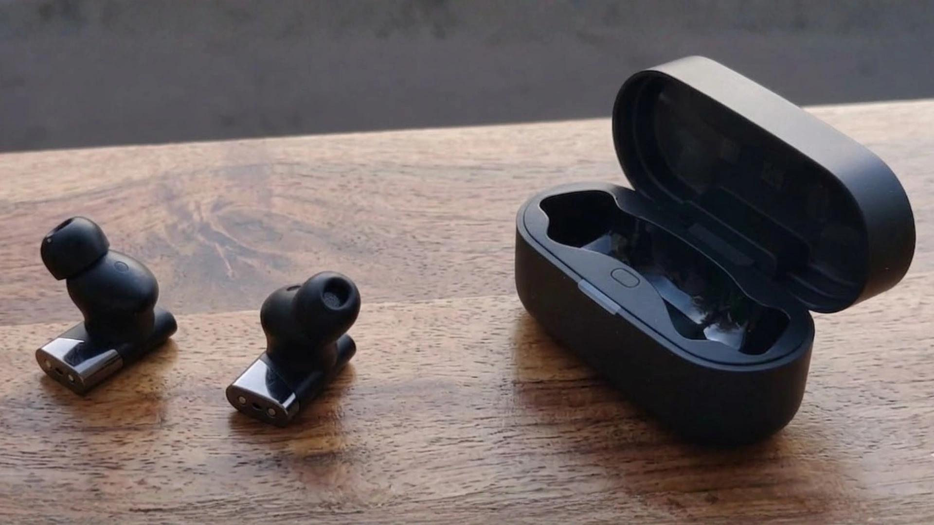 Status Pro X earbuds placed beside open charging case on wooden table showing compact design and magnetic case lid