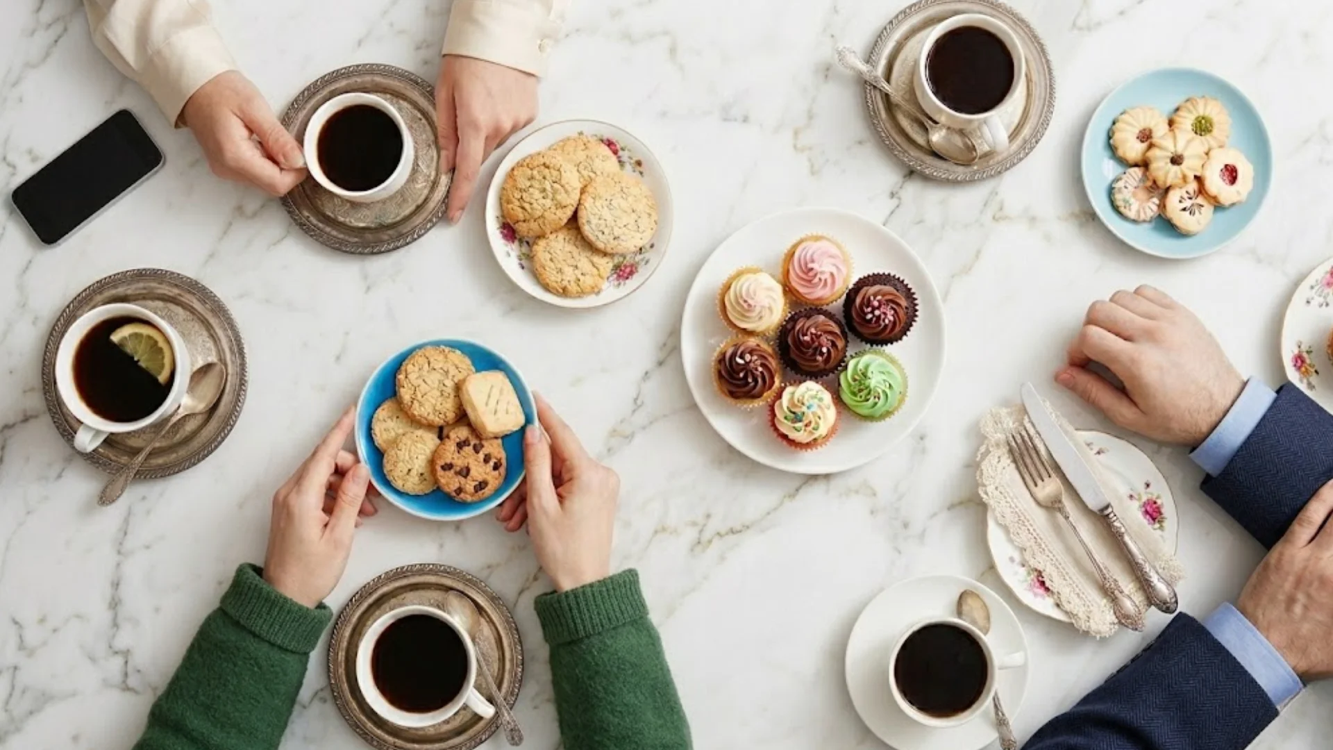 Top view of coffee cups, cookies, and cupcakes on marble table with people sharing dessert and coffee together