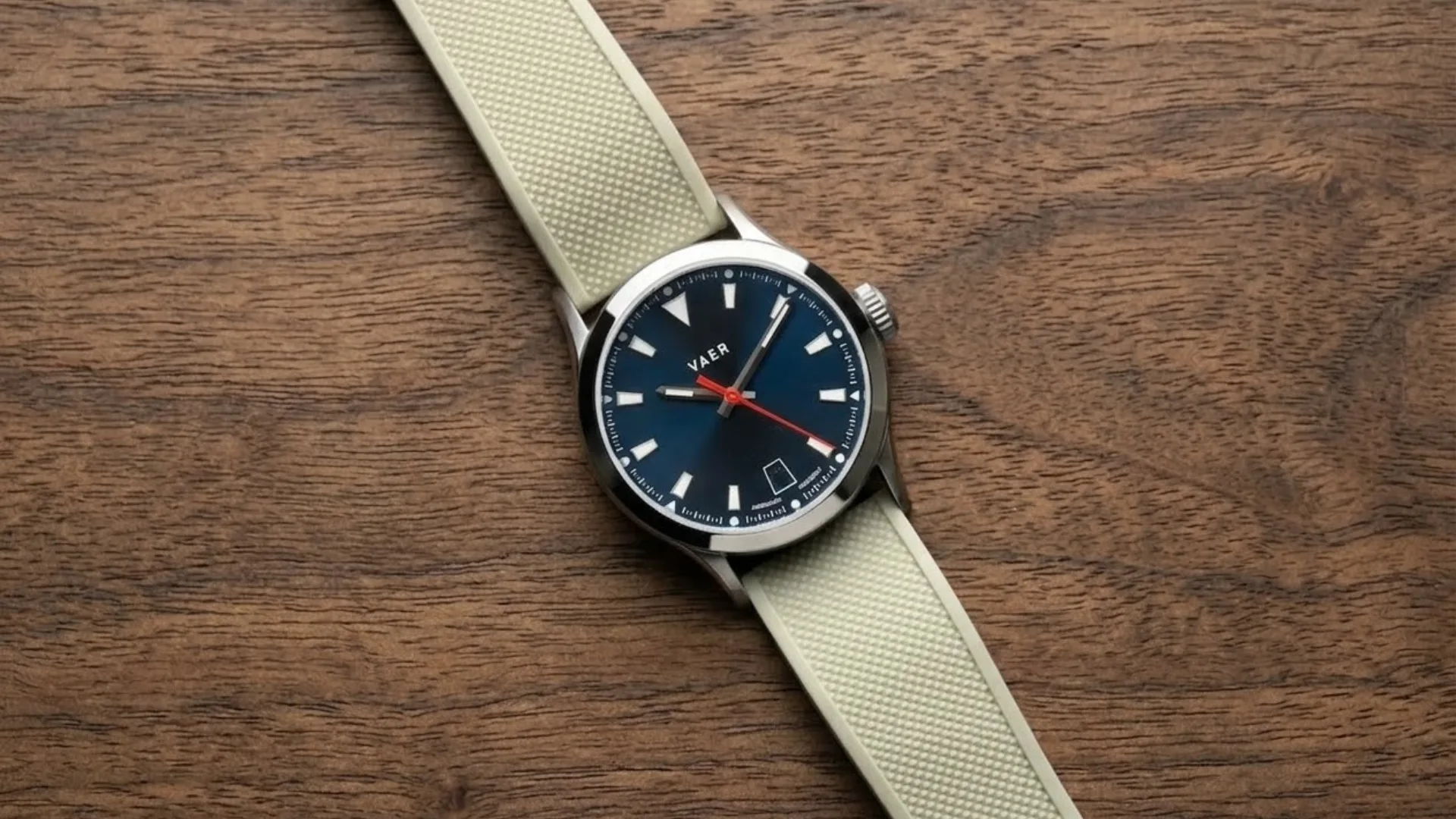 Vaer automatic watch with blue dial, red seconds hand, date window, and beige rubber strap placed on wooden surface