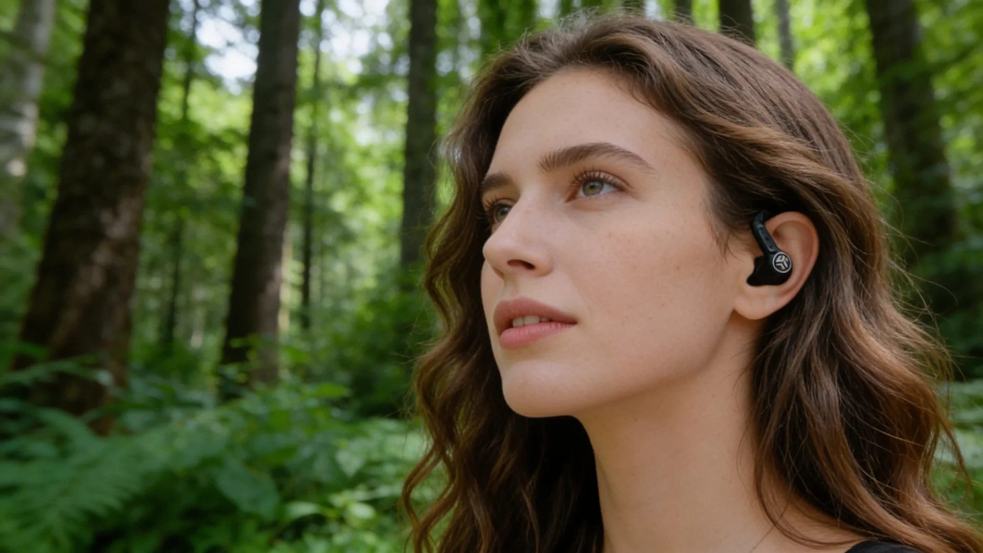 Woman wearing JLab wireless earbuds outdoors in a forest while listening to music during a walk in nature