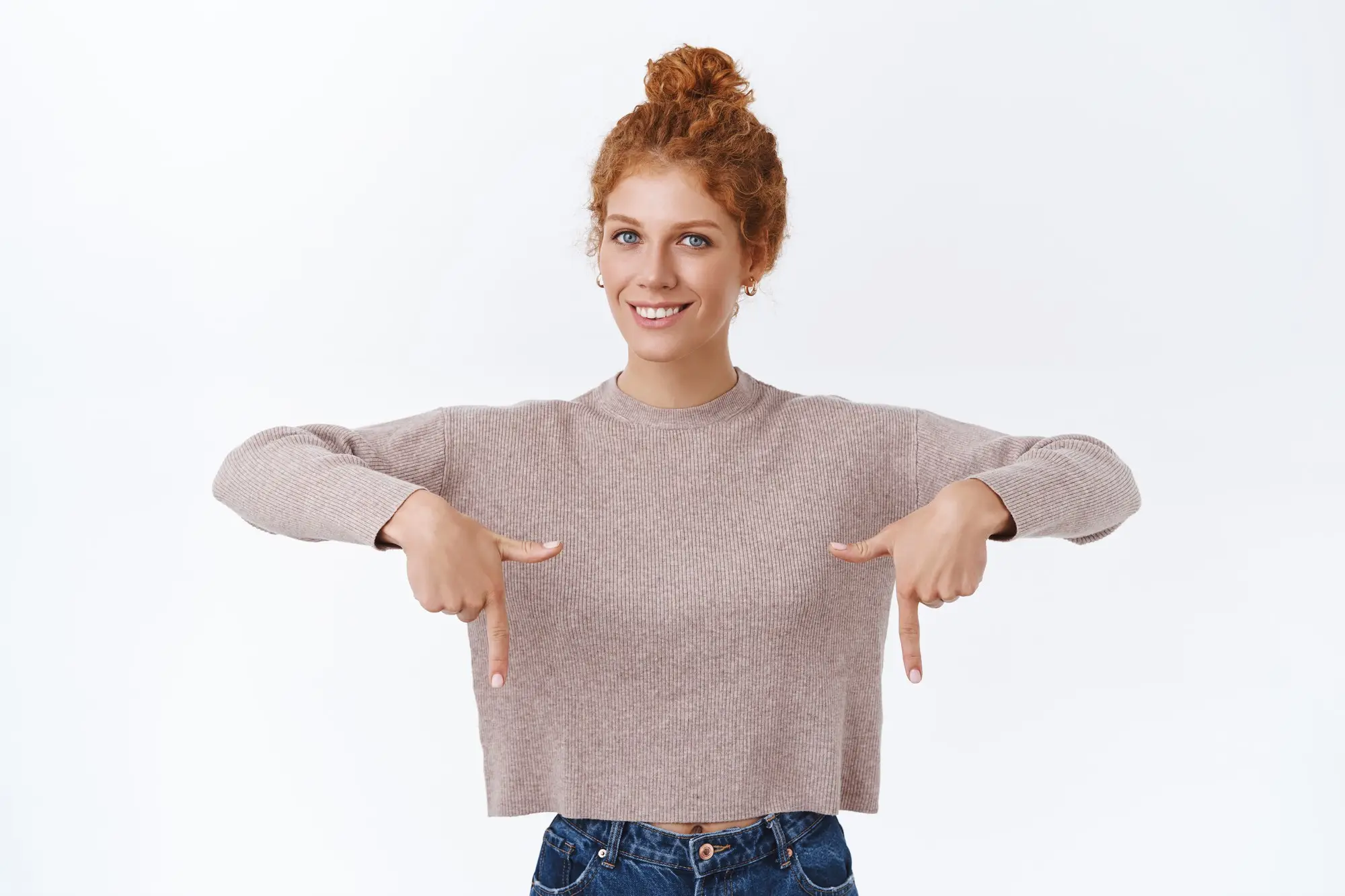 Woman with curly hair pointing downwards in casual beige sweater against white background