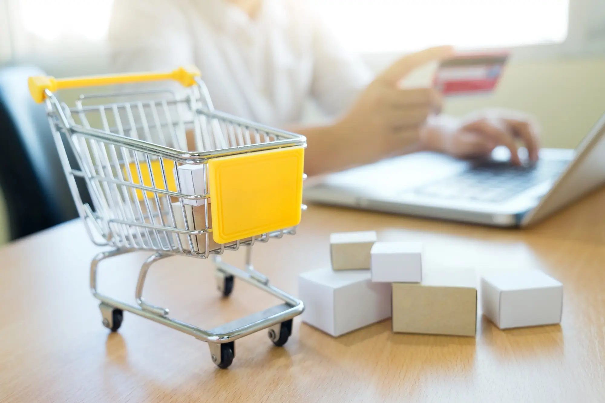 Mini shopping cart and cardboard boxes on desk in front of person using laptop