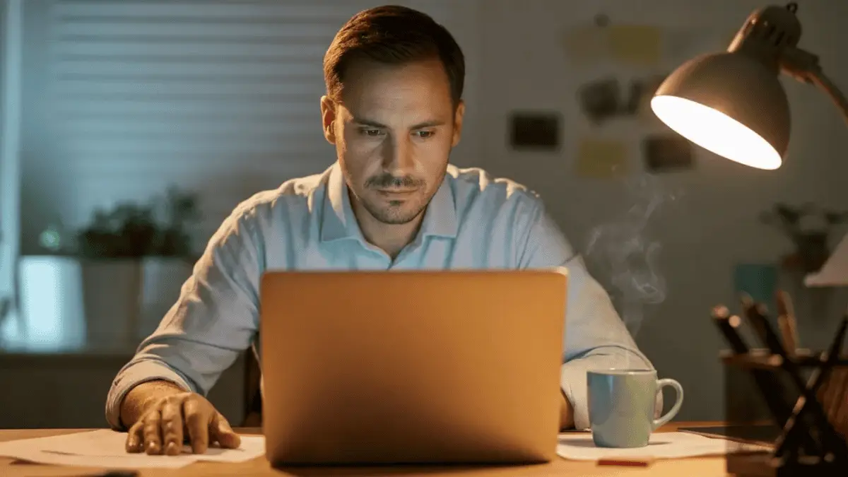 Man working on laptop at desk in dimly lit office with lamp and steaming mug