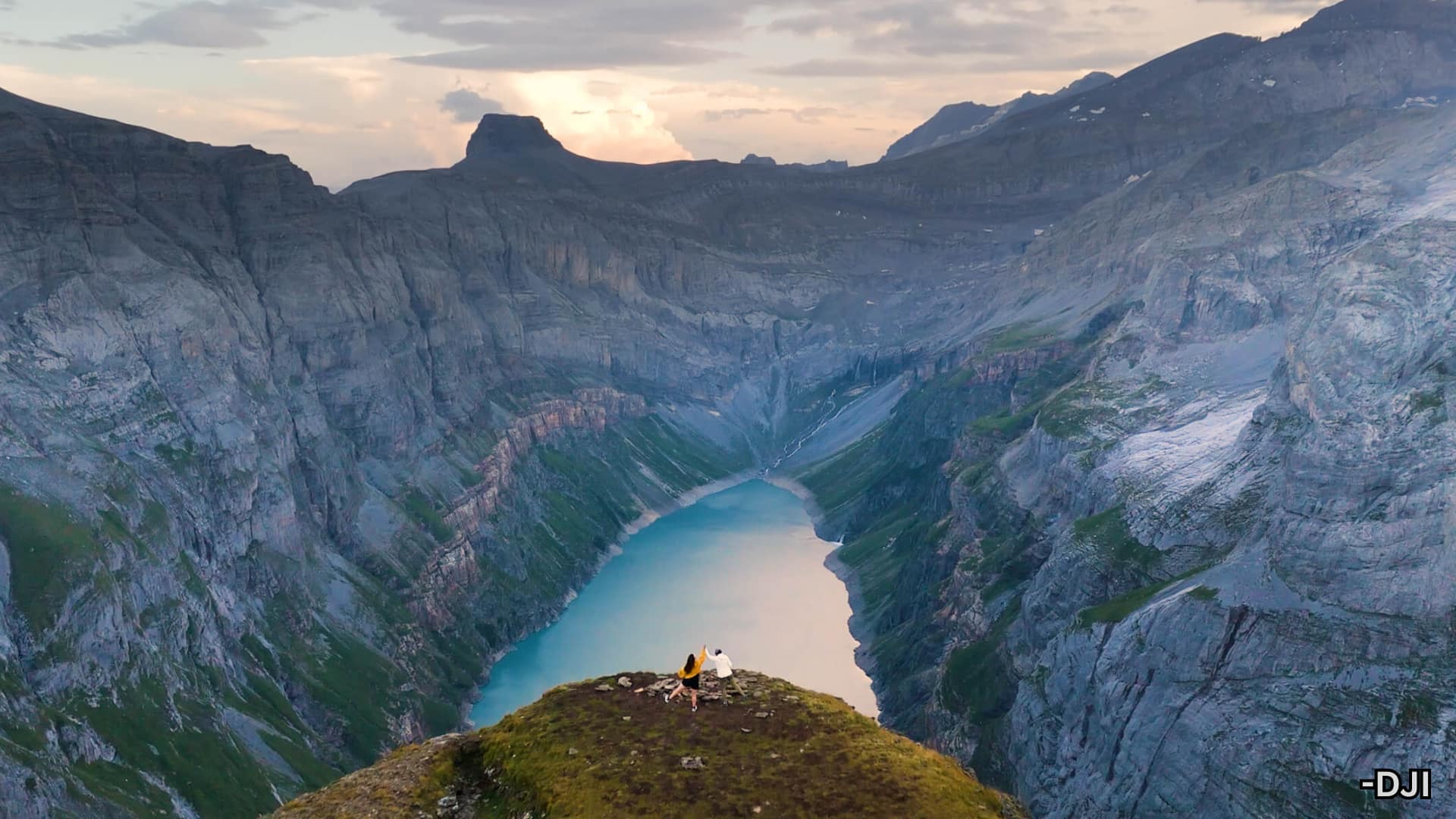 Aerial view of mountain valley with turquoise lake and two people on cliff, captured by DJI Mini 4 Pro drone