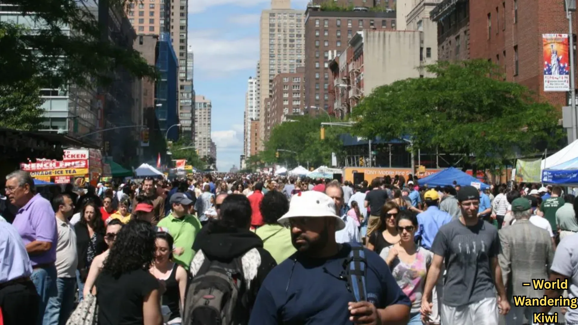 Busy city street fair with large crowd, food stalls, and tents lined between tall buildings on a sunny day