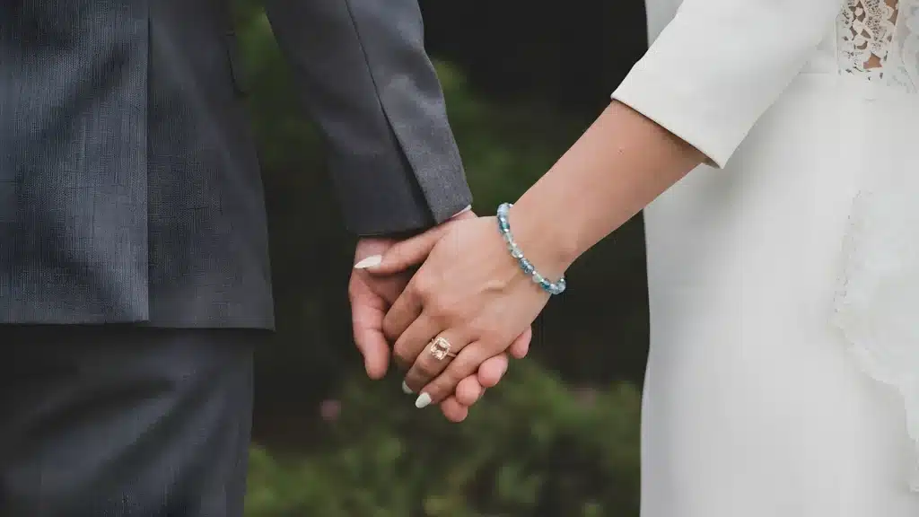 Close up of a bride and groom holding hands outdoors with a visible diamond wedding ring on the bride’s finger