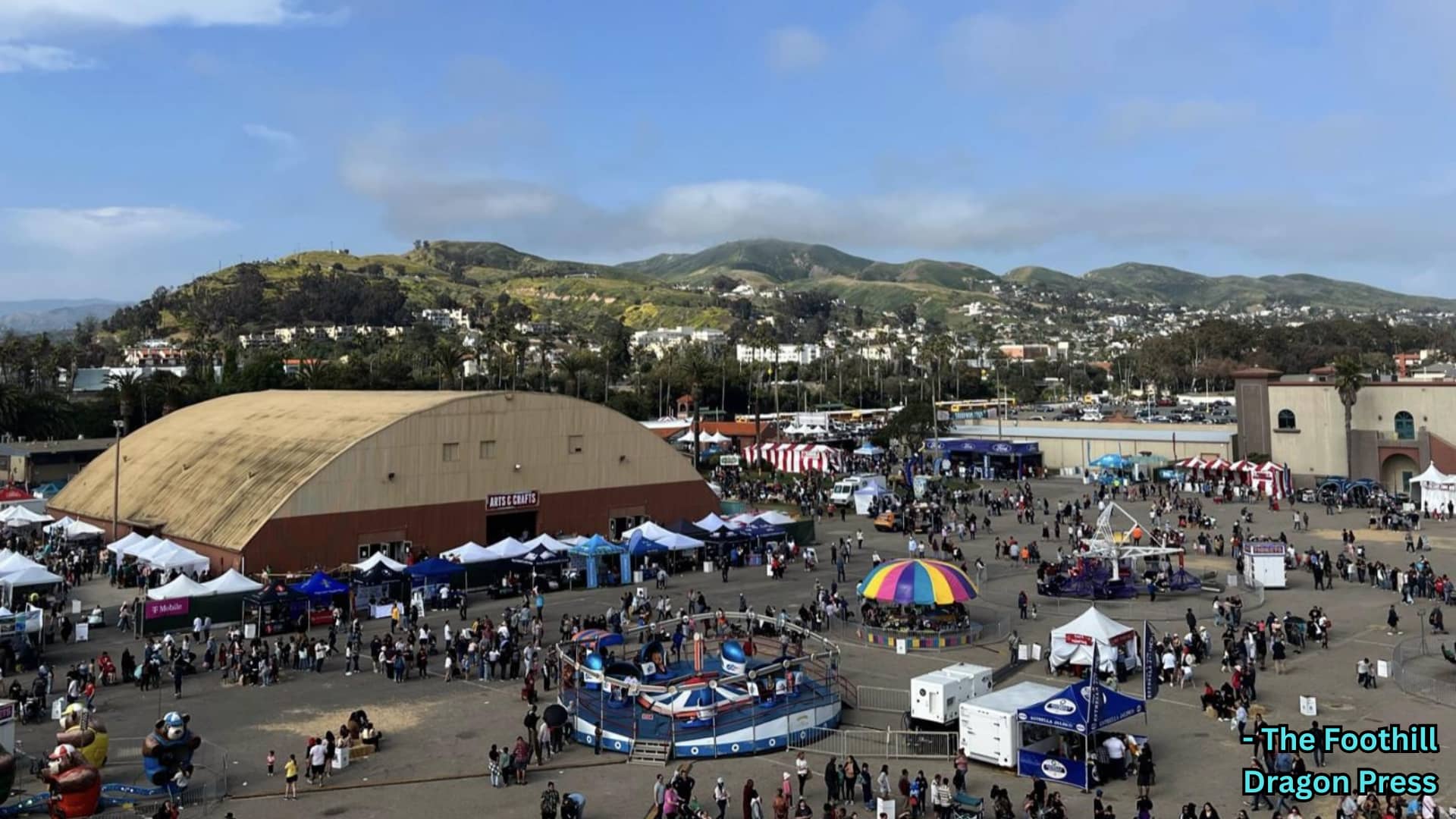 Crowd at outdoor fair with rides, tents, and stalls, set against hills and buildings under a clear sky
