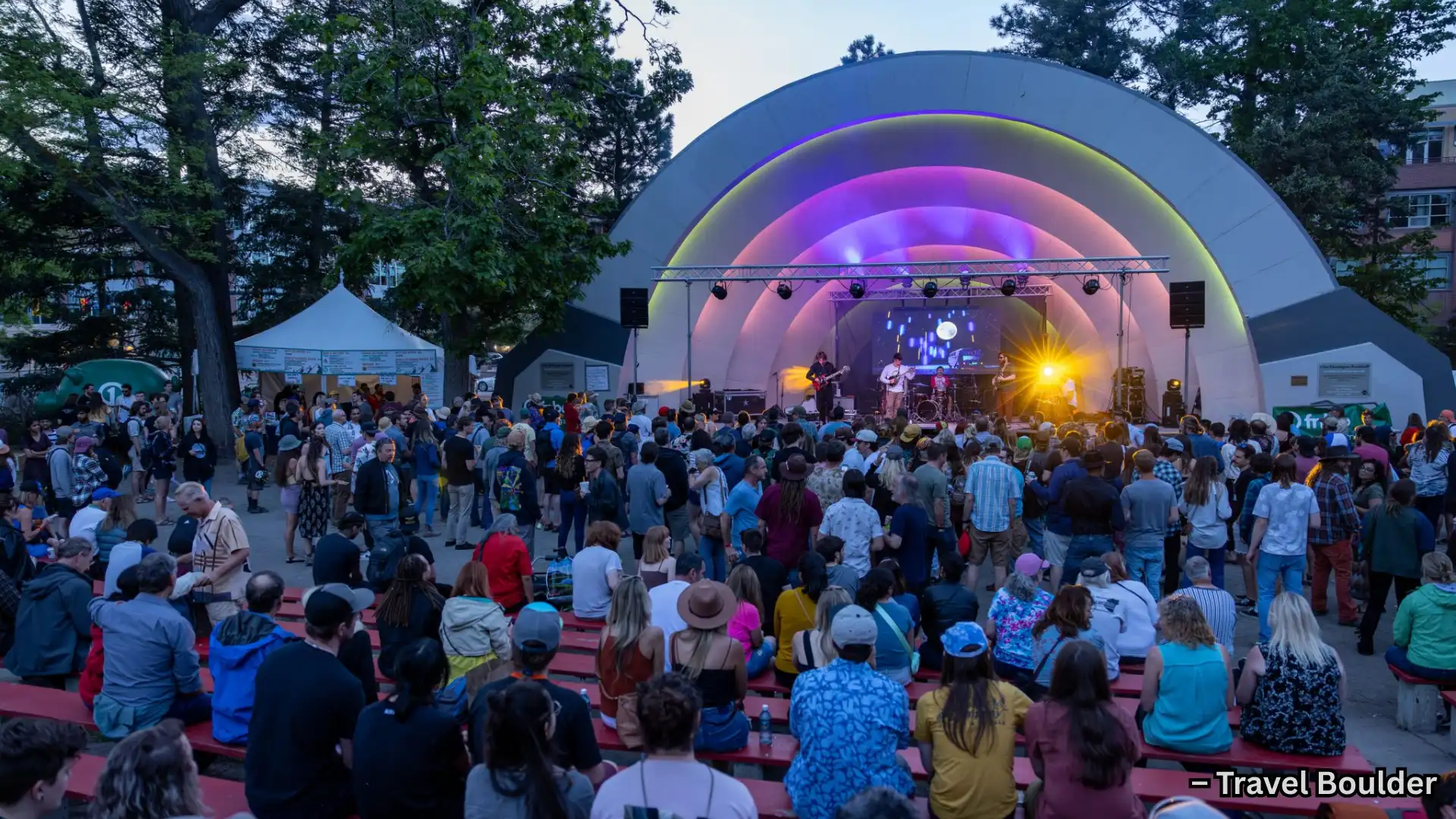 Crowd watching live band at outdoor amphitheater, colorful lit stage and evening festival atmosphere