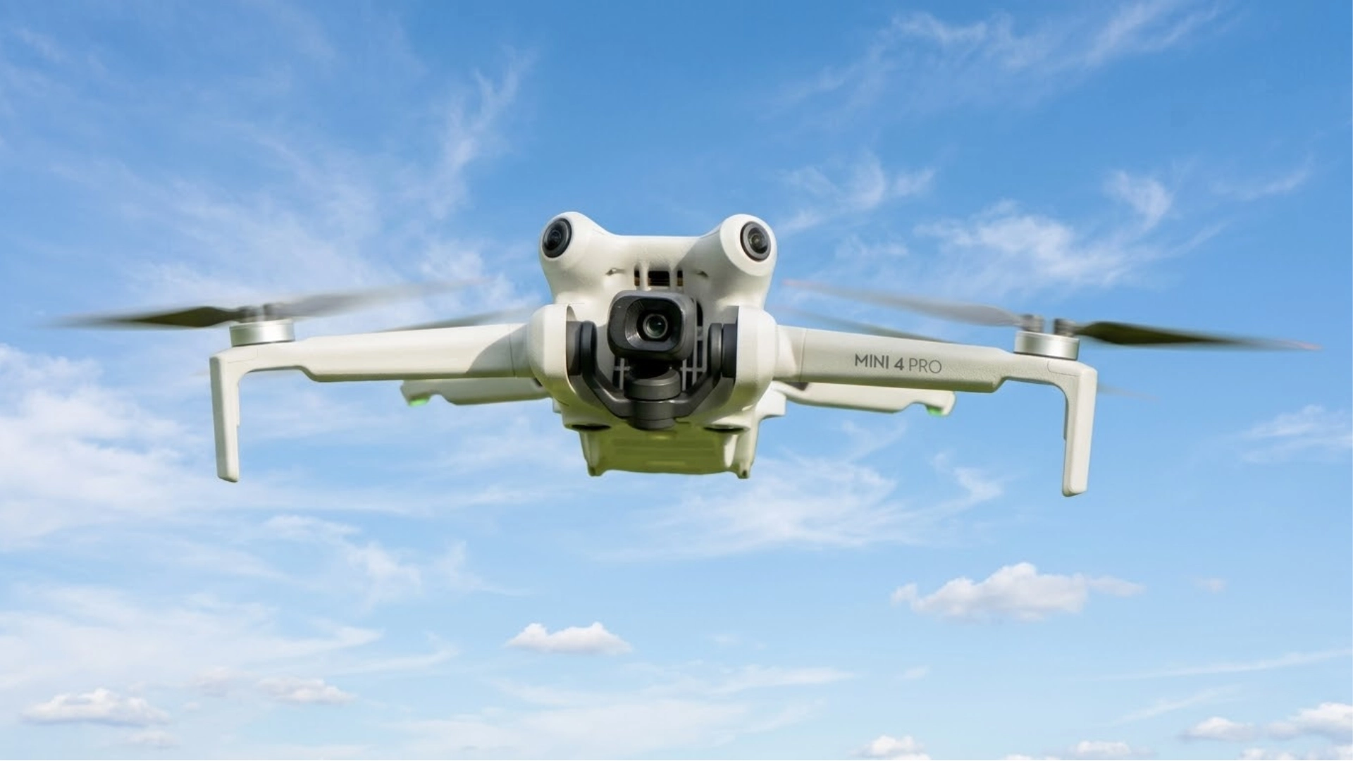 DJI Mini 4 Pro drone flying mid air against blue sky with clouds, showing front camera and propellers in motion