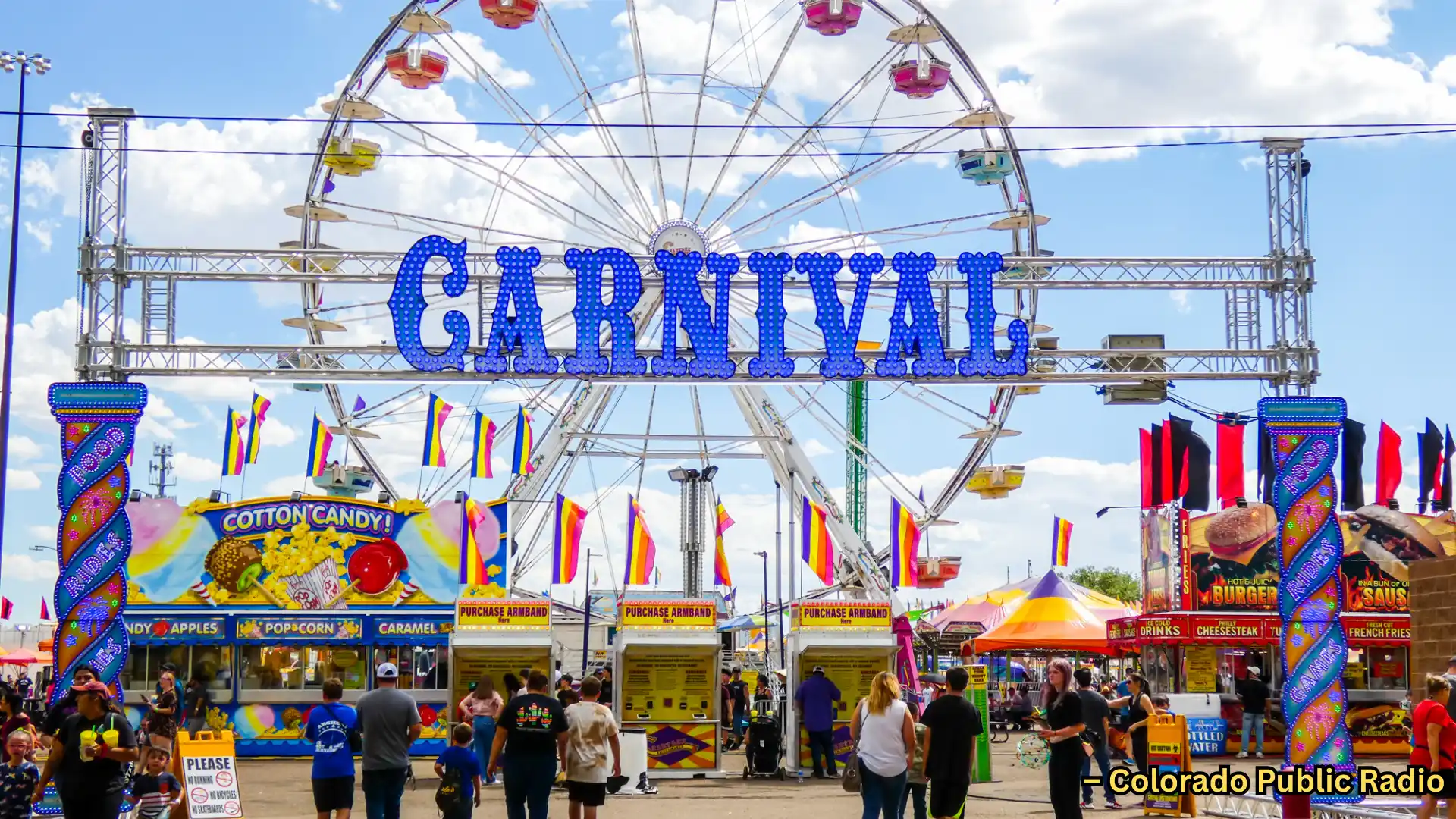 Entrance to carnival with Ferris wheel, colorful stalls, and people walking under bright daytime sky