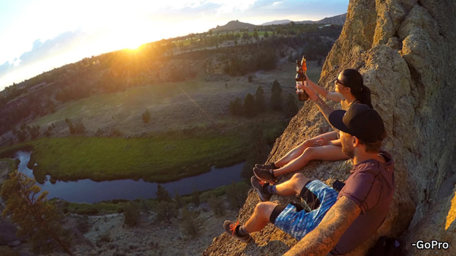 GoPro wide angle shot of couple sitting on cliff at sunset with river valley view and dramatic landscape in background