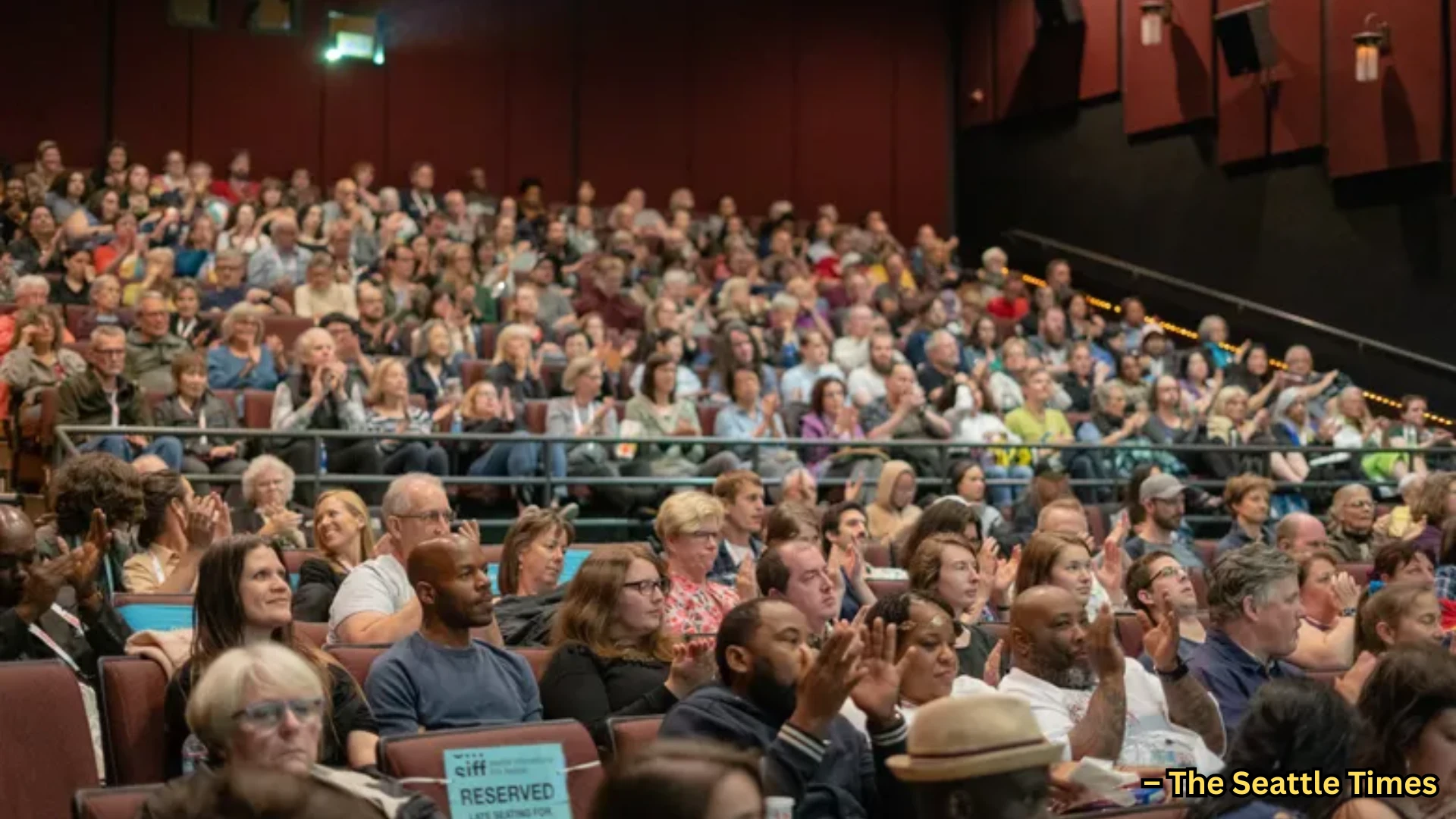 Large audience seated in theater, people clapping and watching stage during indoor event or presentation