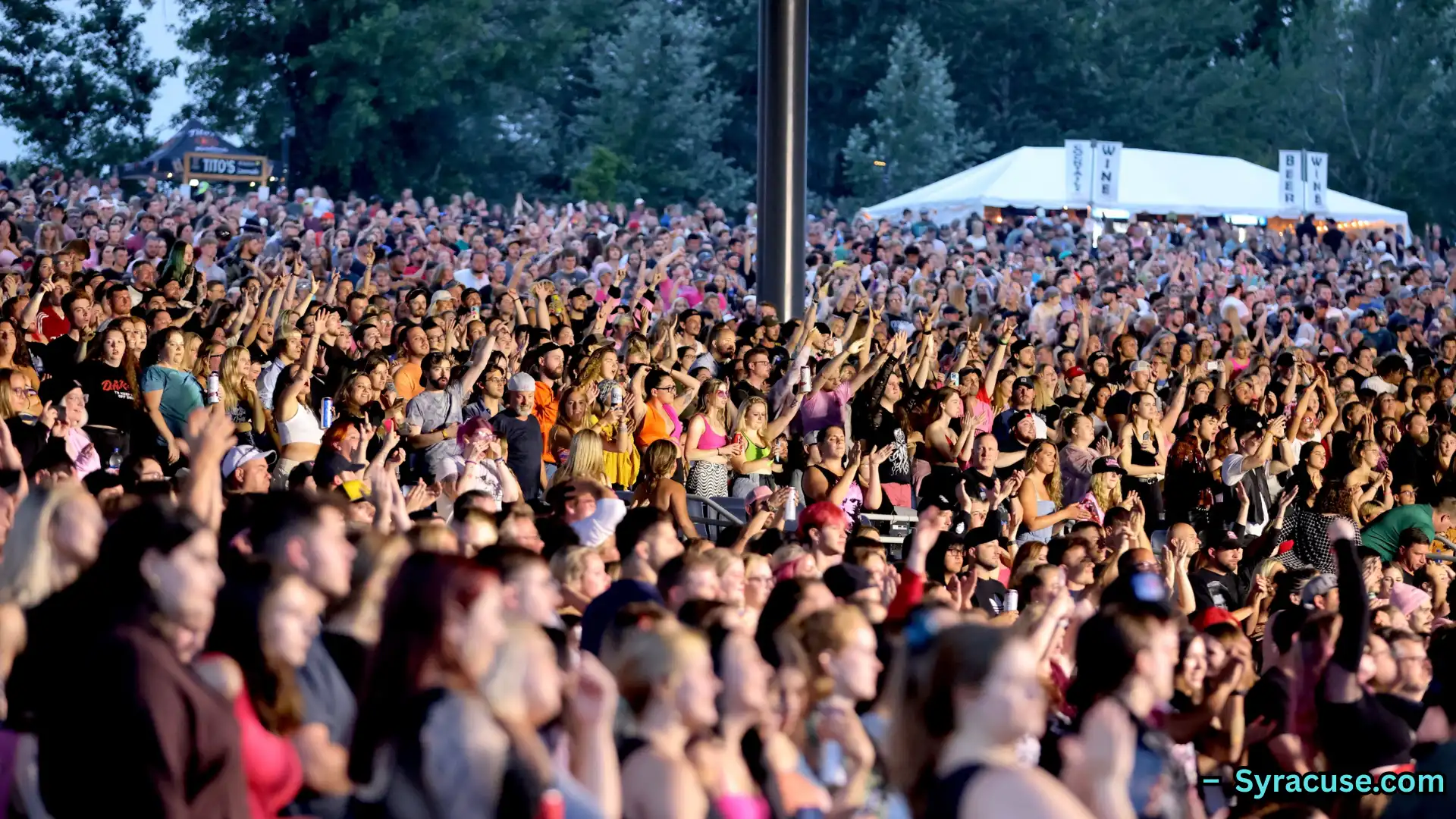 Large crowd cheering at outdoor concert, people raising hands and enjoying live music in evening setting