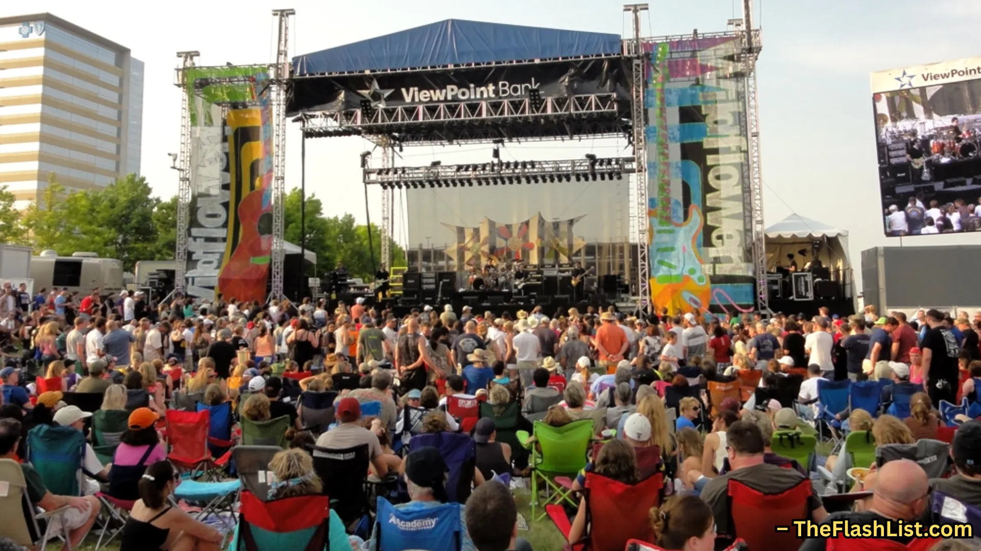 Large crowd watching live band on outdoor stage at music festival, people seated and standing in open venue