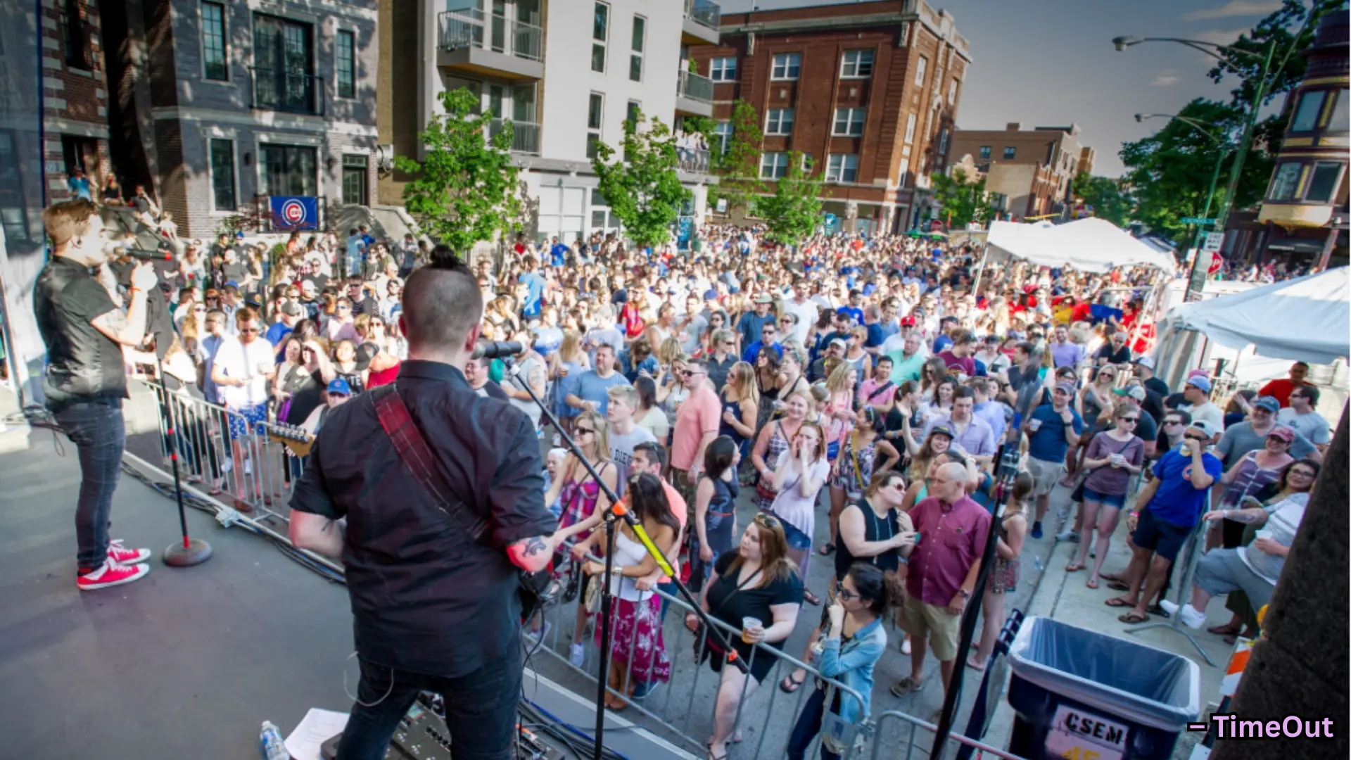 Live band performing on outdoor street stage, large crowd gathered in city setting during daytime event