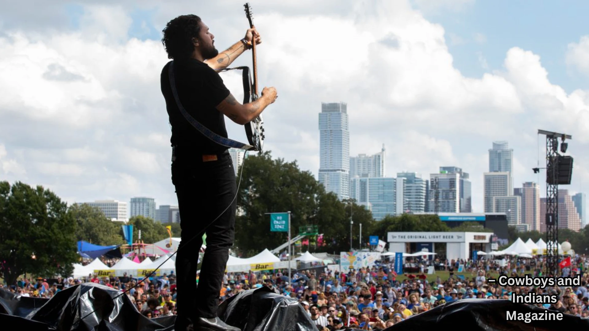 Musician playing guitar on outdoor stage before large crowd, city skyline visible under cloudy sky