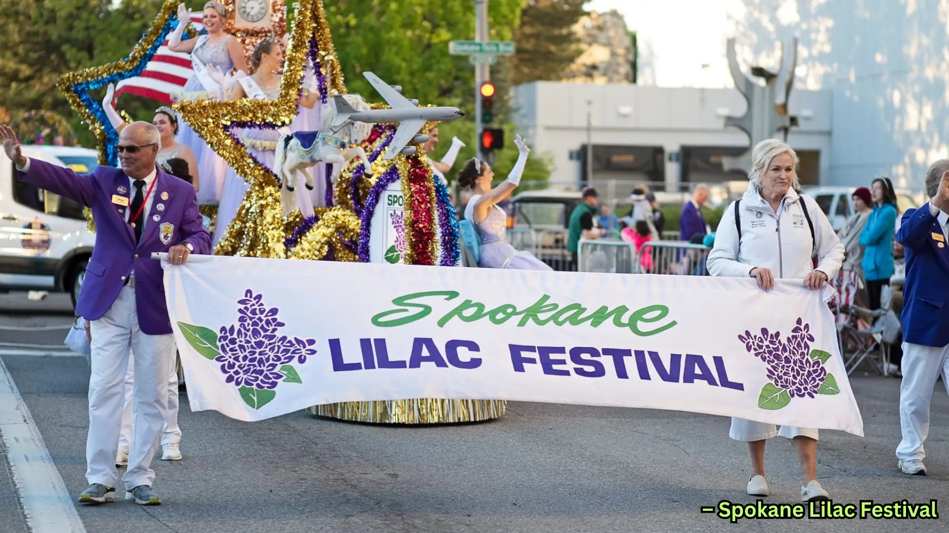 Parade for Spokane Lilac Festival with decorated float, banner carriers, and performers in festive costumes on street