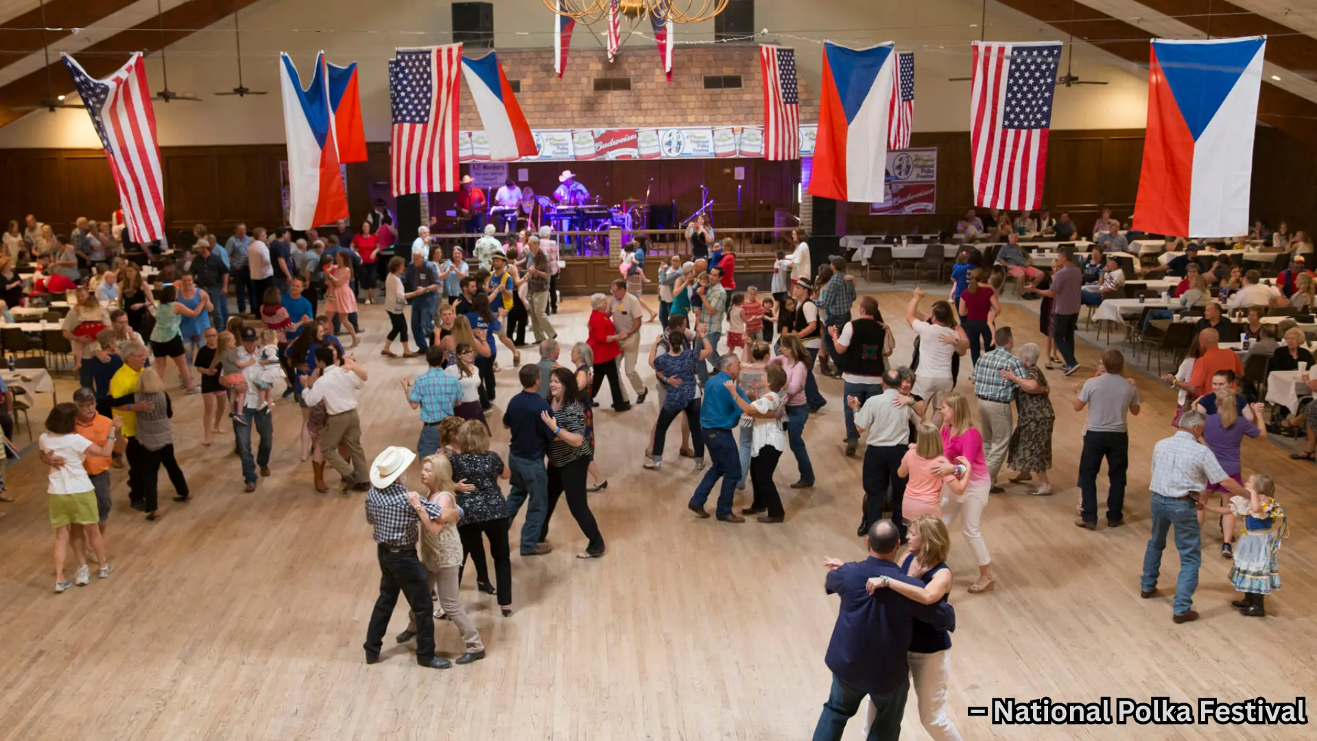 People dancing at indoor community hall event with live band, flags hanging, and large crowd enjoying social gathering