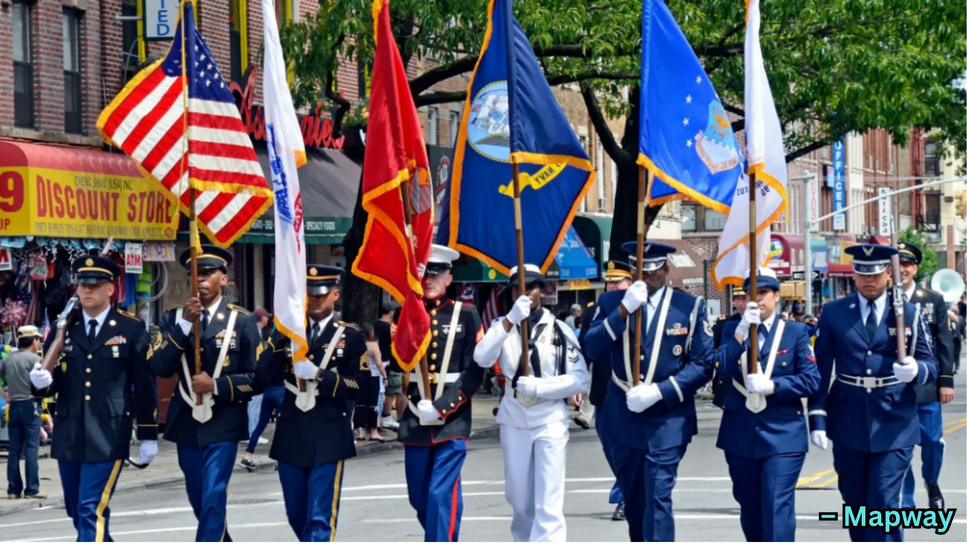 Uniformed service members march in parade carrying flags through city street with spectators and shops in background