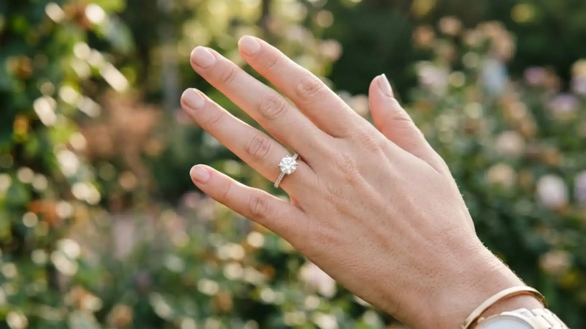 Woman’s hand showing diamond engagement ring outdoors with blurred garden background in natural light highlighting the ring