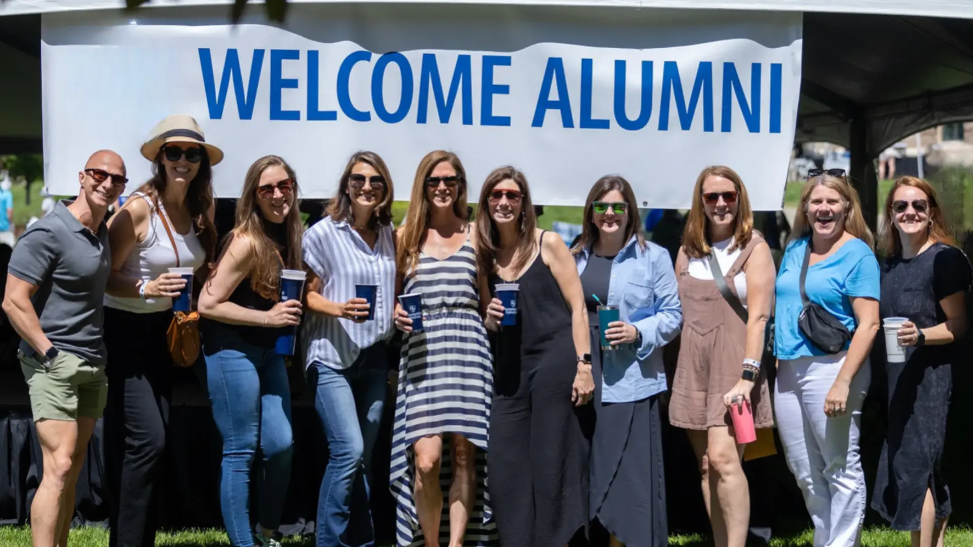 Alumni event group smiling under welcome banner, holding drinks and posing together outdoors
