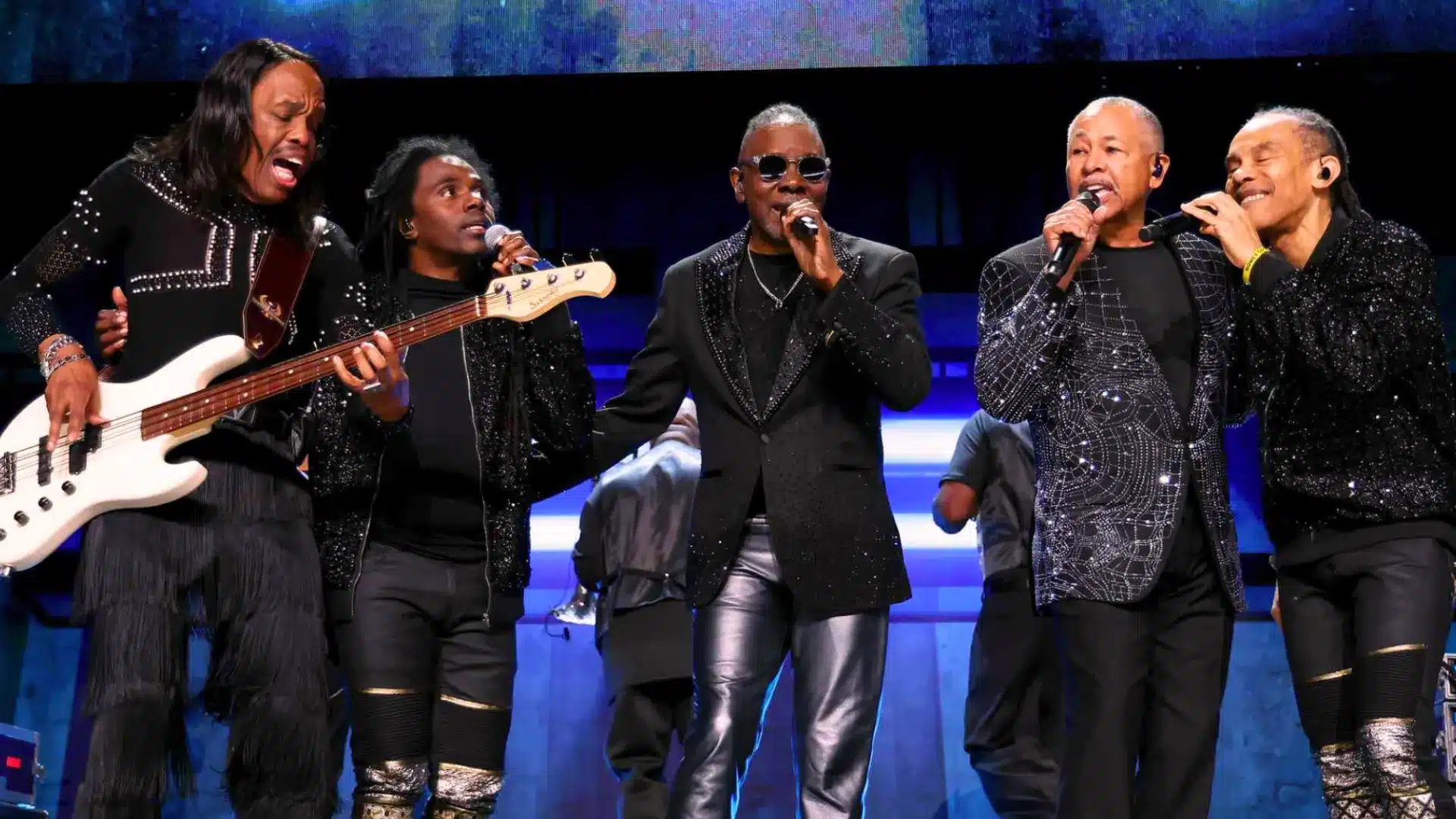 Benefit concert with singers performing live on stage, holding microphones and playing instruments under bright lights