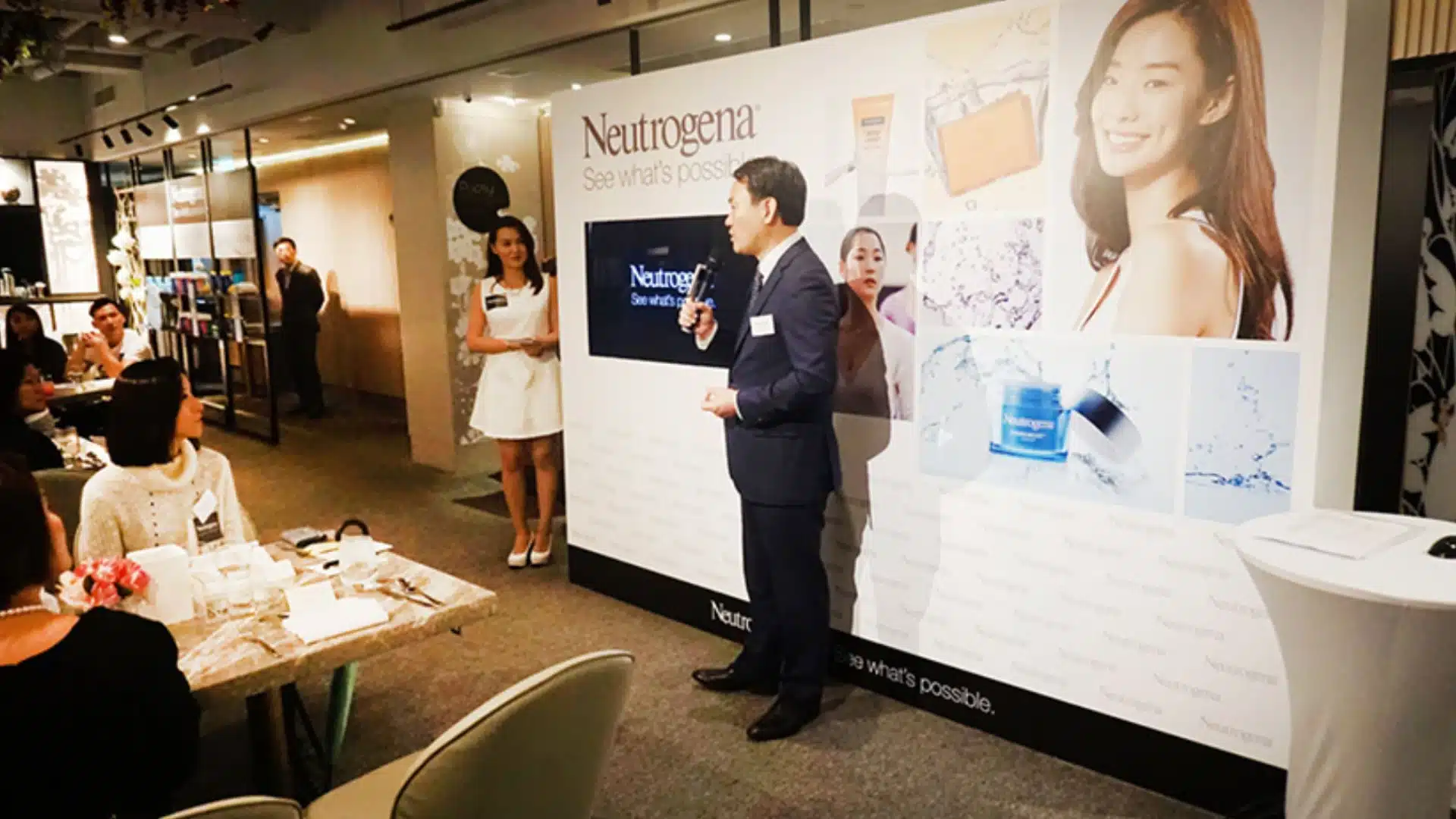 Corporate product launch event with speaker presenting brand display to seated audience in indoor setting