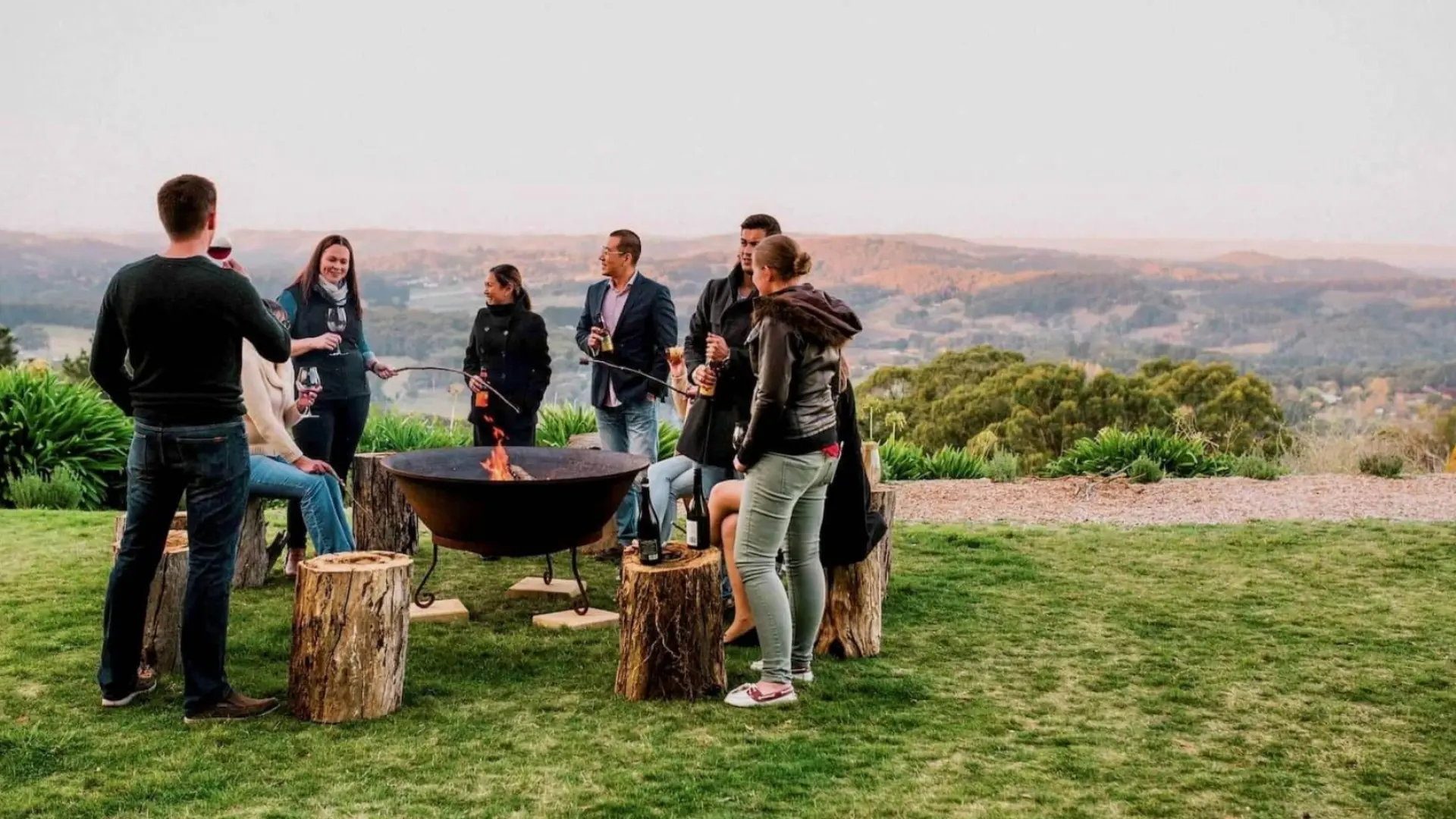Corporate retreat group socializing around fire pit outdoors with scenic hills in background