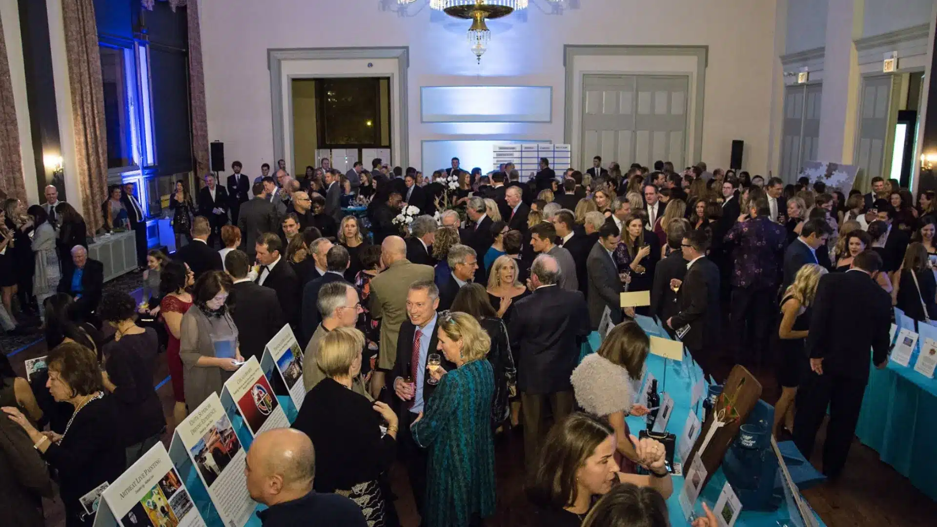 Crowded fundraising auction event indoors with guests mingling, viewing items, and socializing in formal setting