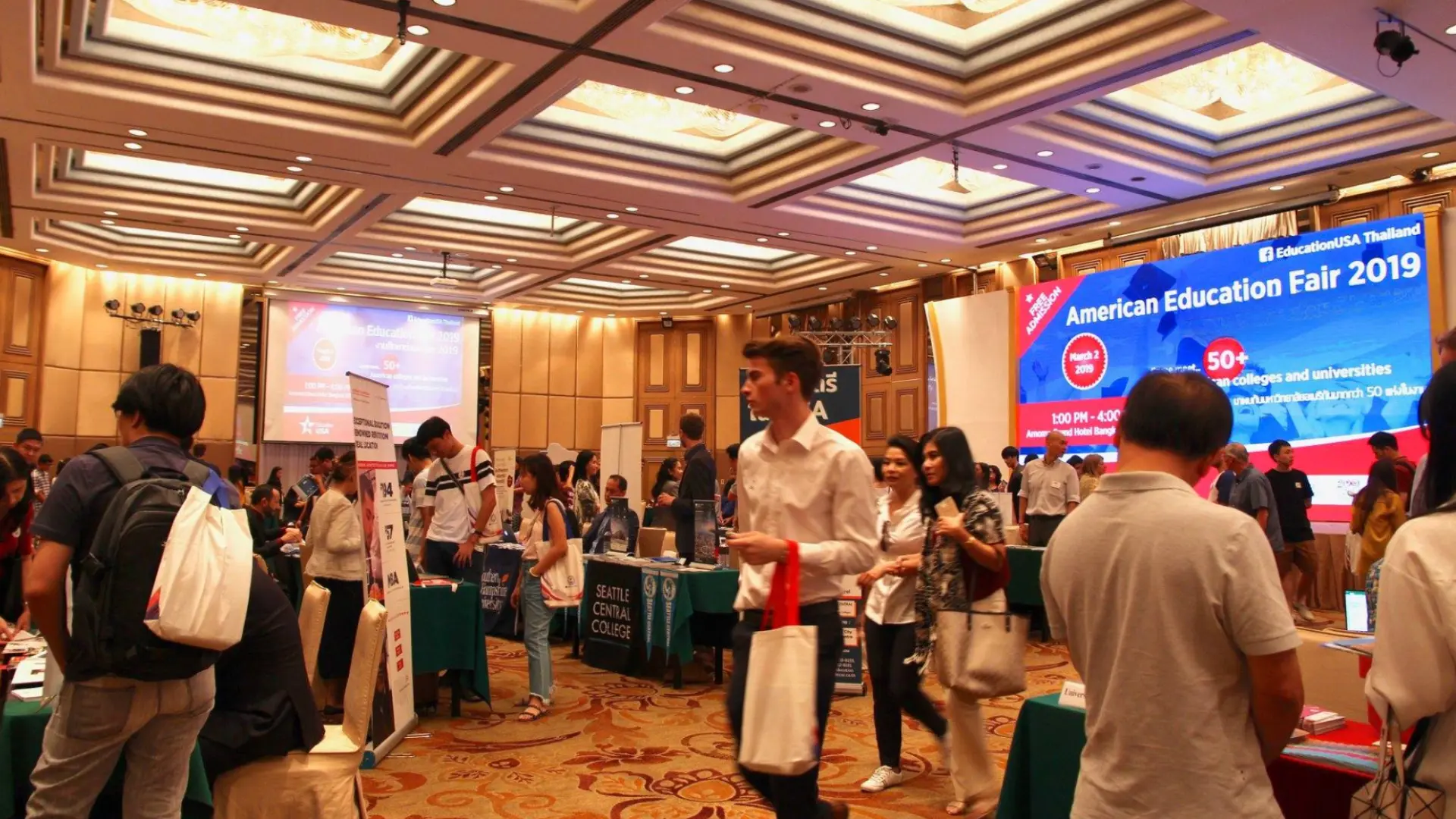 Educational fair with booths and students exploring colleges, walking through busy indoor exhibition hall