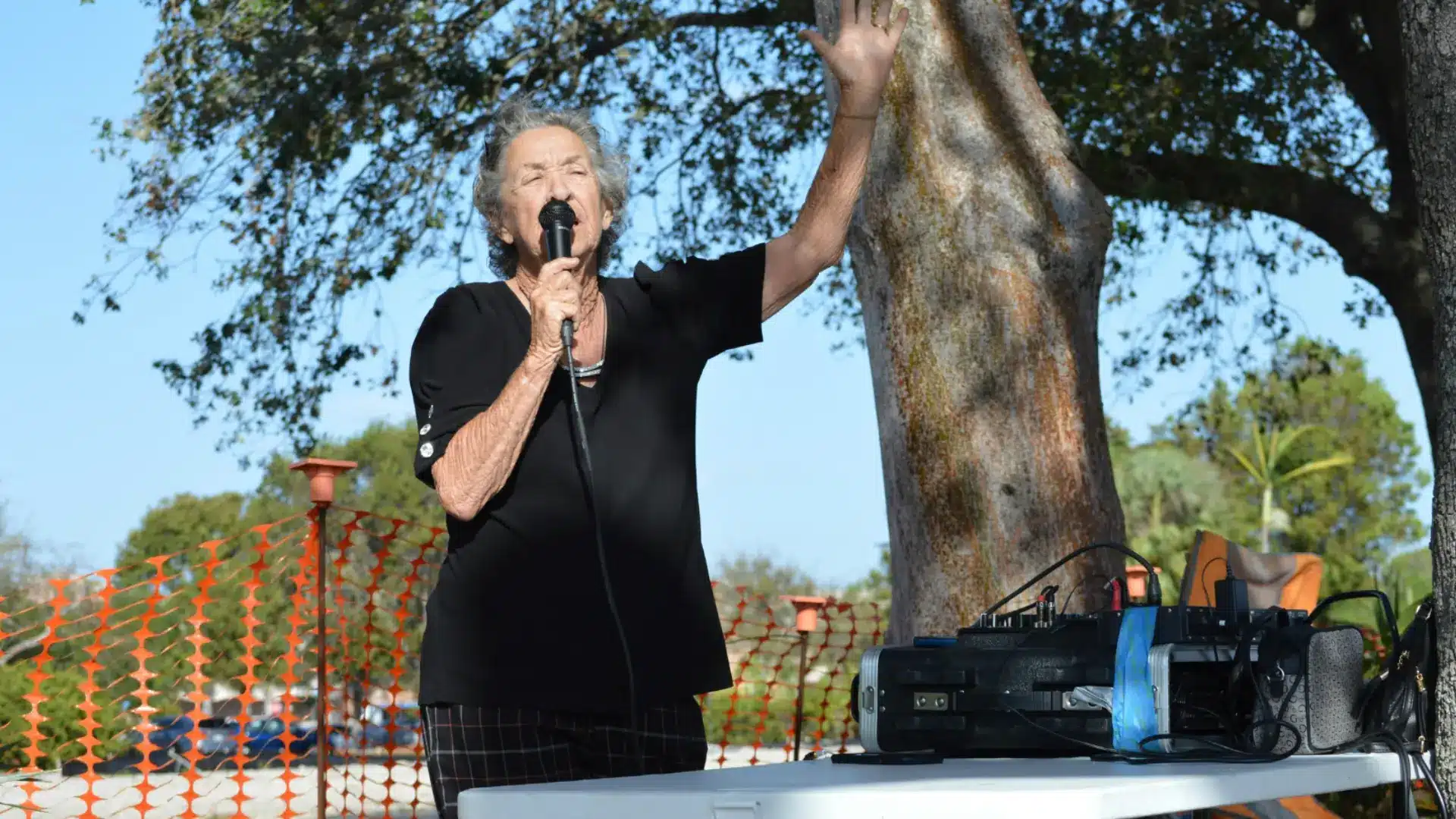 Elderly woman speaking into microphone outdoors during community charity event with audio setup