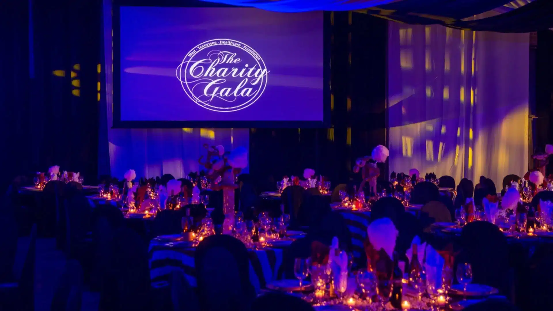 Elegant charity gala setup with decorated tables, candles, and large screen displaying event branding in dim lighting
