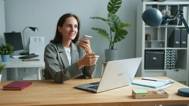 Businesswoman in grey blazer using smartphone with laptop at wooden desk in modern office
